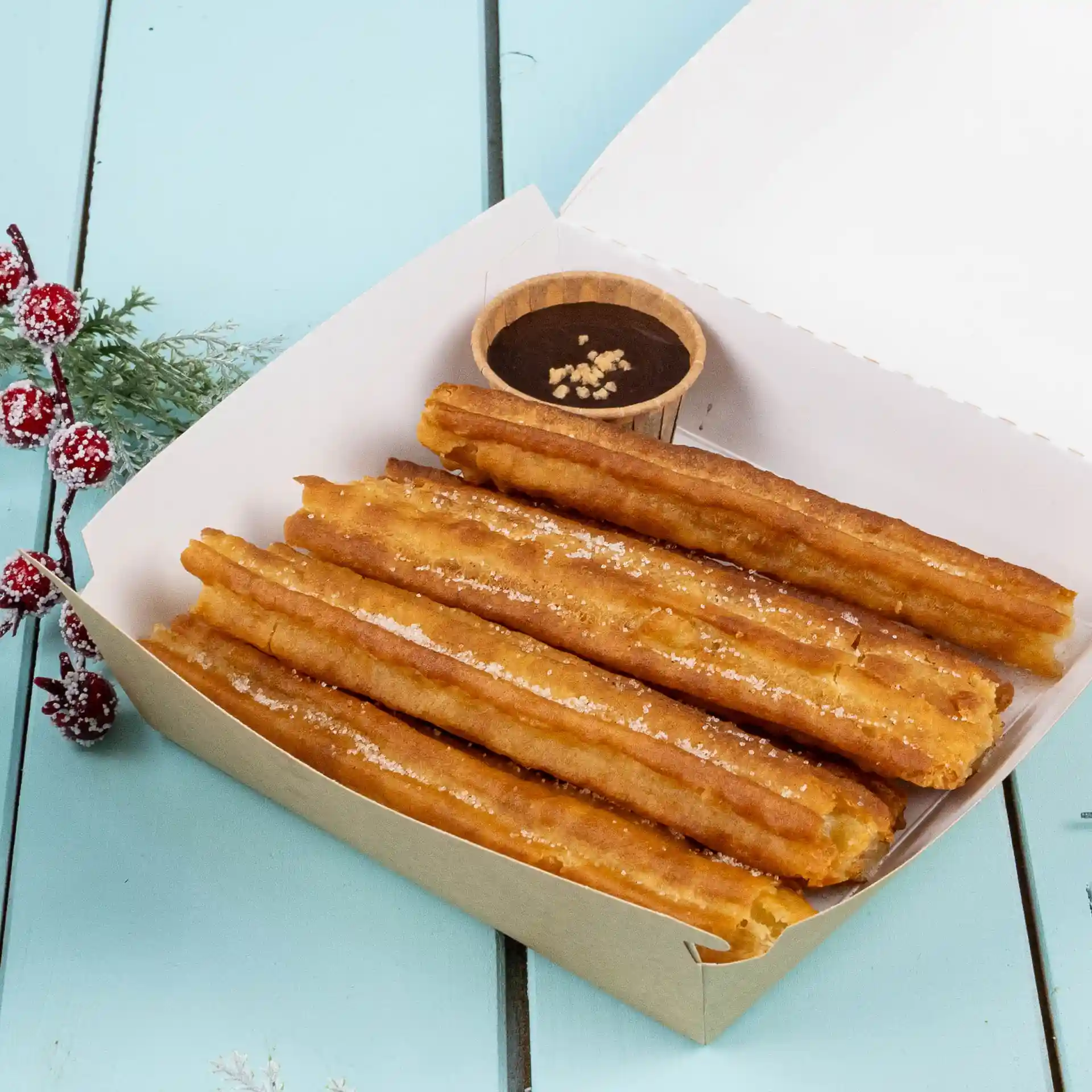 Churros in una scatola di carta con una piccola tazza marrone di salsa al cioccolato su una superficie blu.