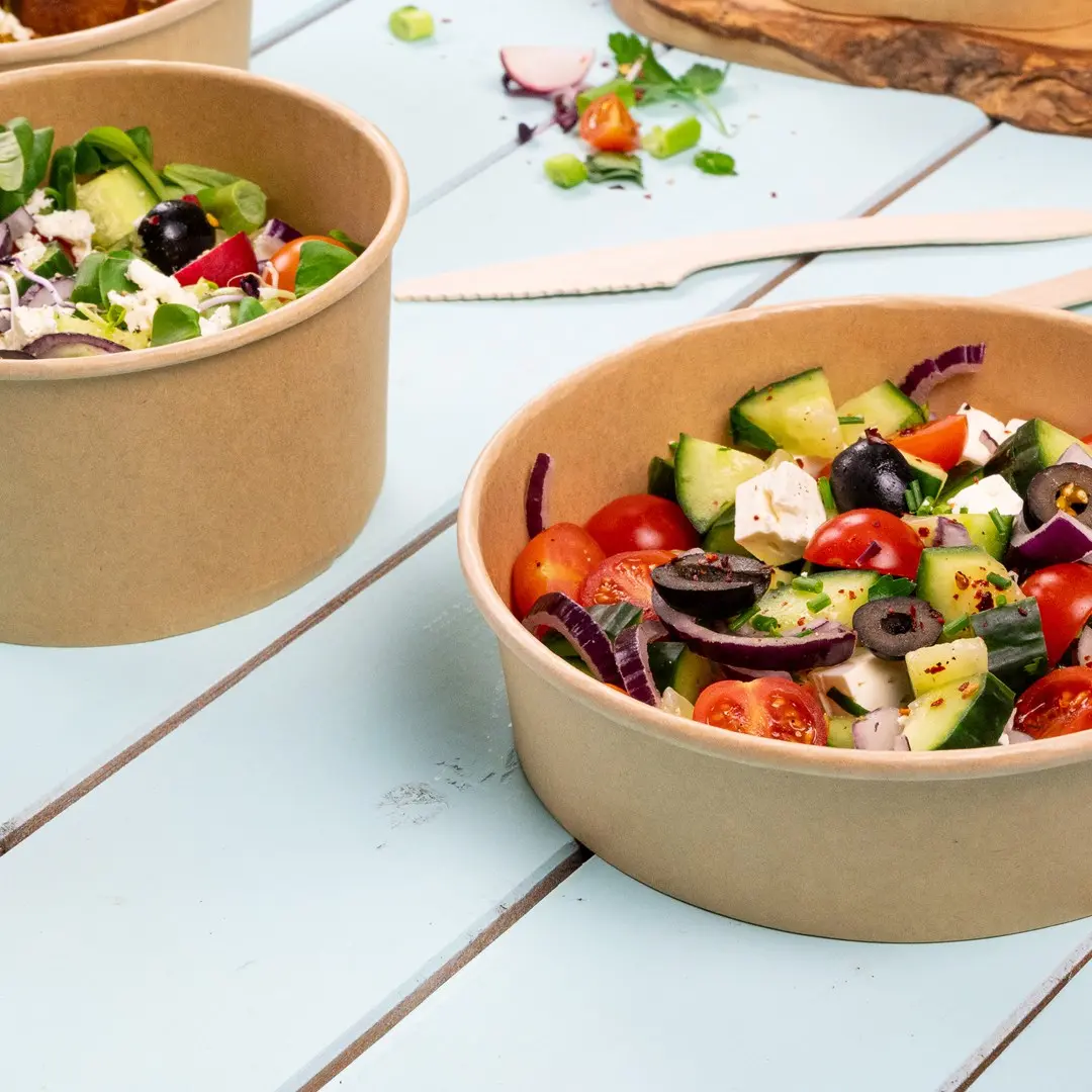Due ciotole di carta marrone piene di verdure miste, tra cui pomodori, cetrioli, olive e verdure, su una superficie in legno blu.