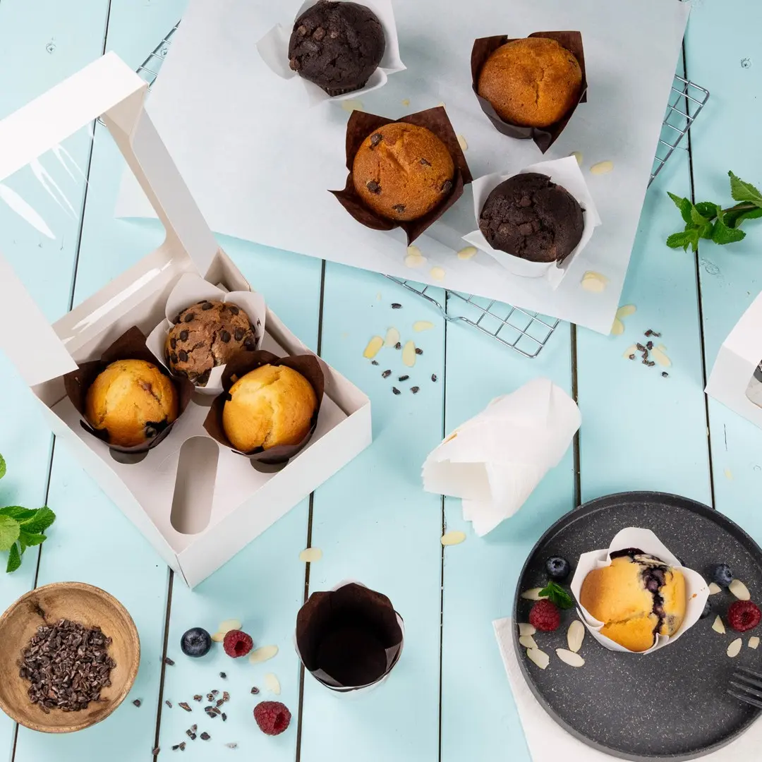 Scatola bianca contenente tre muffin, circondata da muffin extra, bacche e ingredienti da forno su una superficie blu.