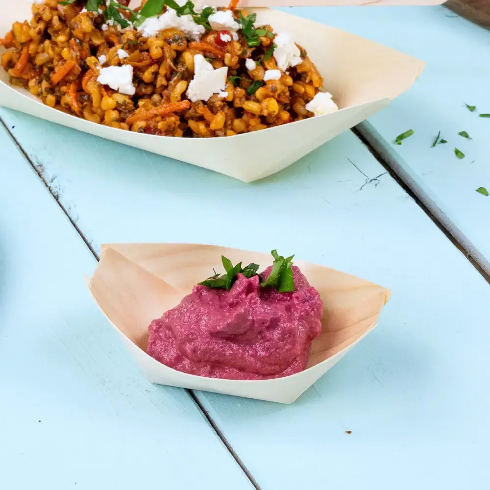 Piatto a forma di barca in legno con salsa rosa guarnita con erbe verdi, accanto a un piatto più grande di cereali e verdure miste.
