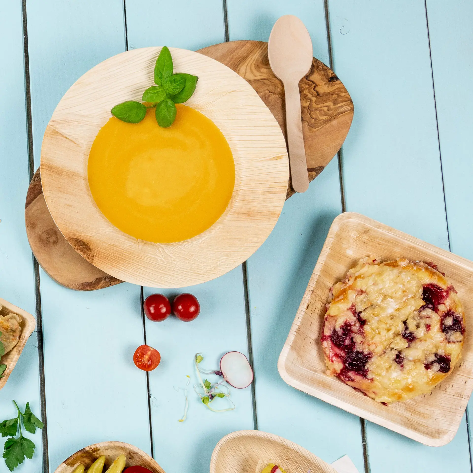 Un piatto rotondo profondo in foglia di palma con zuppa gialla e foglie di basilico su un tagliere di legno; alimenti aggiuntivi e un cucchiaio di legno nelle vicinanze.