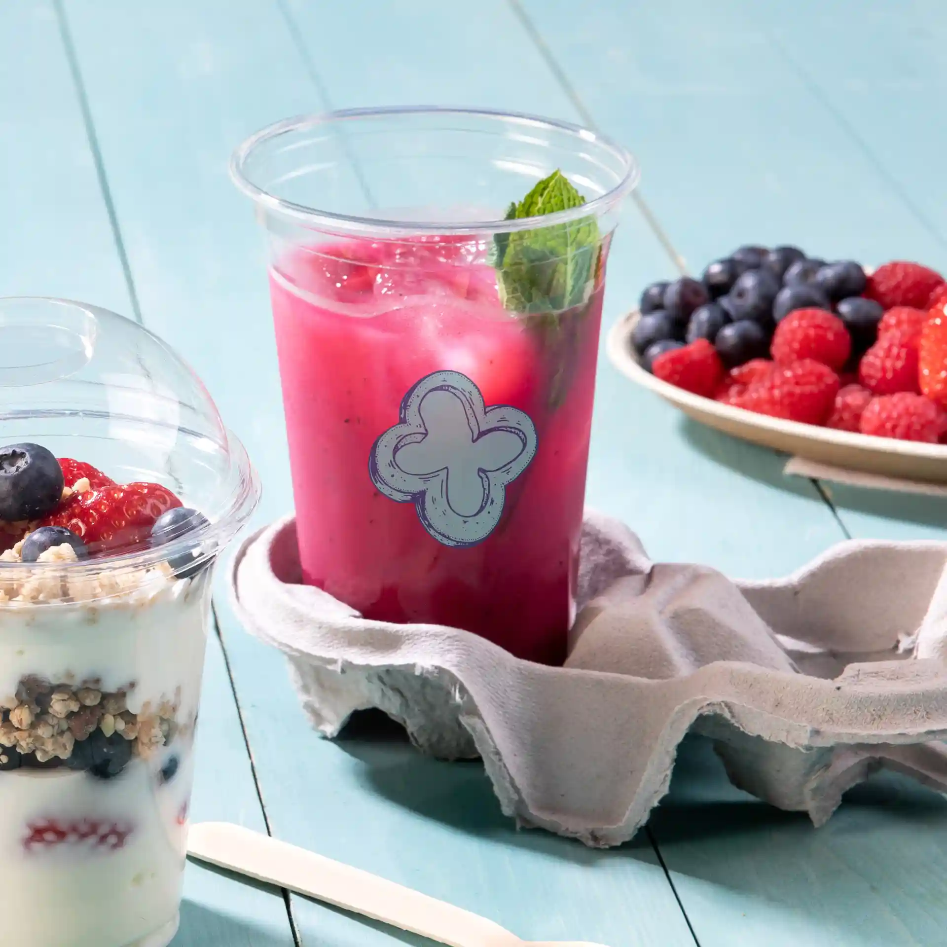 Bicchiere rPET trasparente da 20 oz riempito di bevanda rosa e menta, posizionato in un cartone per uova grigio, con un parfait di yogurt e frutti di bosco nelle vicinanze.