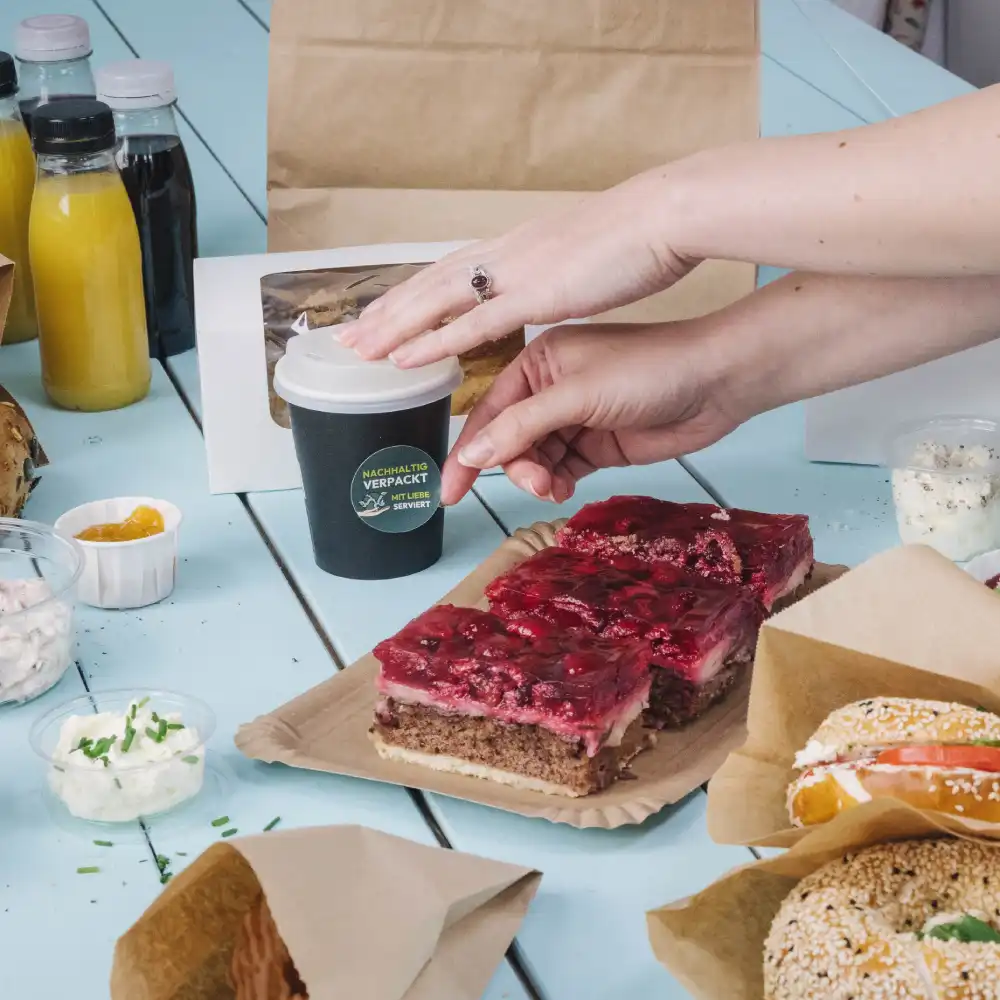 Una mano raggiunge una piccola tazza di caffè nera su un tavolo con vari alimenti; torta a strati, salse e bevande nelle vicinanze.