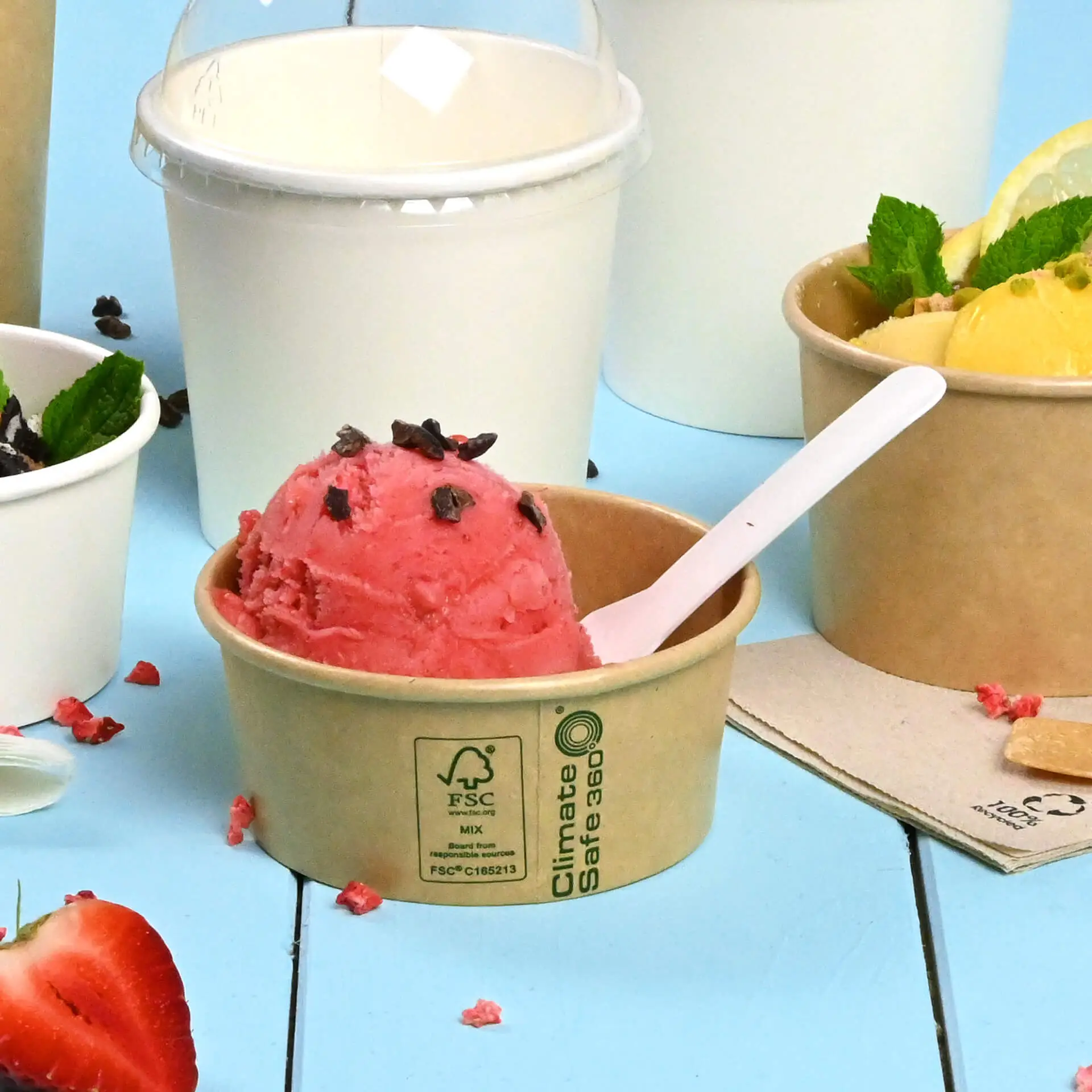 Una coppetta di carta marrone con gelato alla fragola rosa e codette di cioccolato, circondata da altri contenitori per gelato.