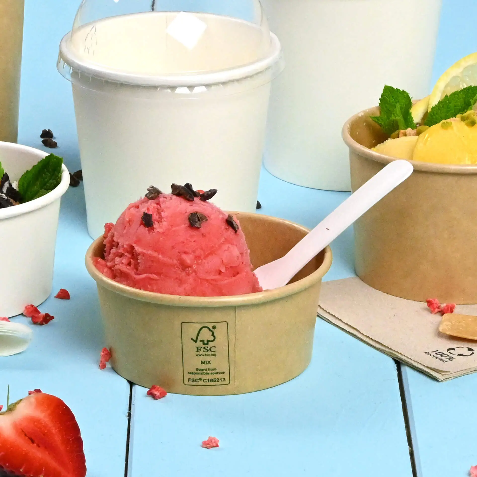 Una coppetta di carta marrone con gelato alla fragola rosa e codette di cioccolato, circondata da altri contenitori per gelato.