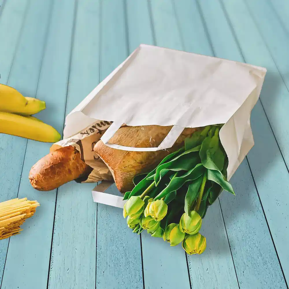 Sacchetto di carta bianco pieno di generi alimentari, tra cui pane, tulipani e banane su una superficie in legno blu.