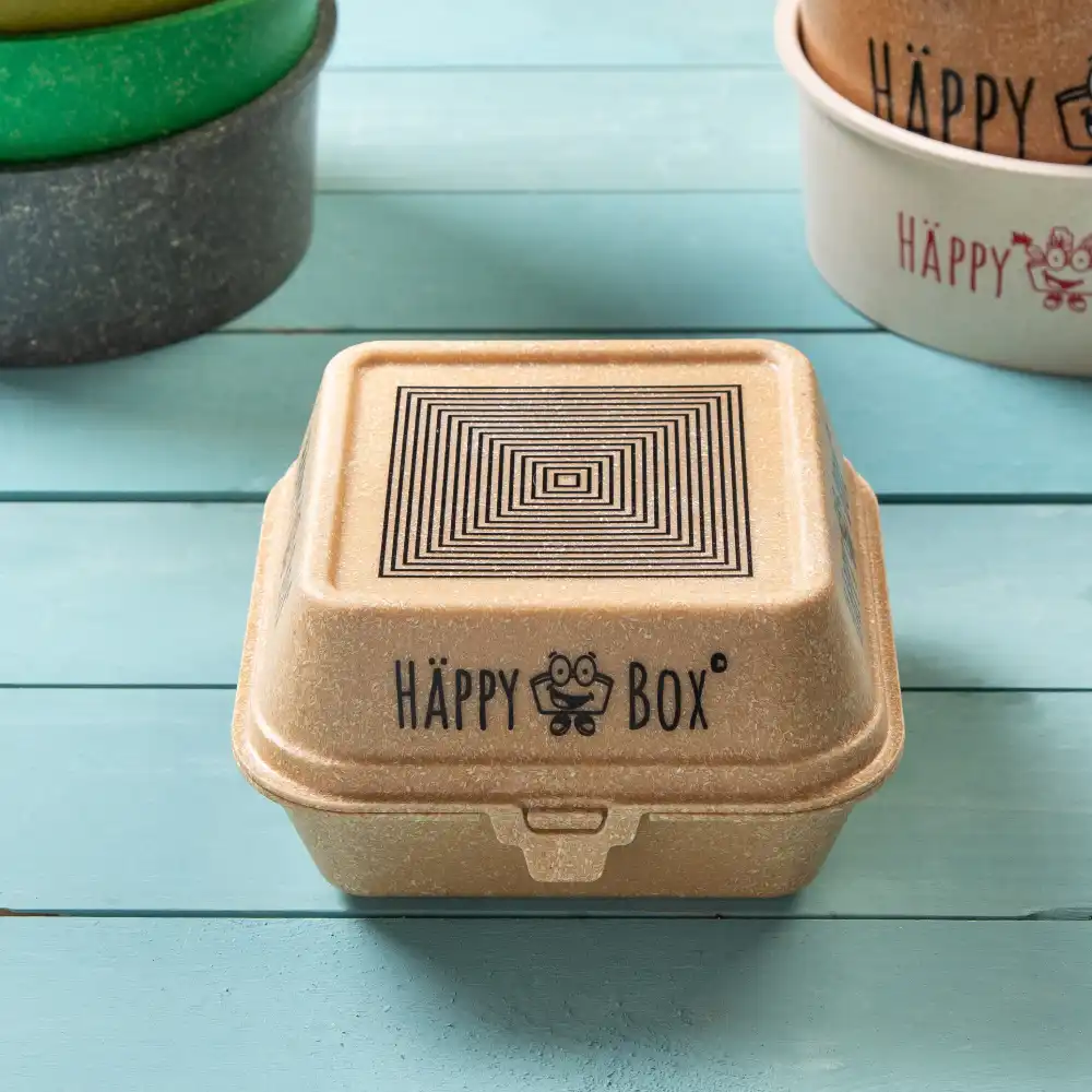 Contenitore alimentare vegano quadrato con un design geometrico nero sul coperchio e il logo "HÄPPY BOX" su una superficie di legno.