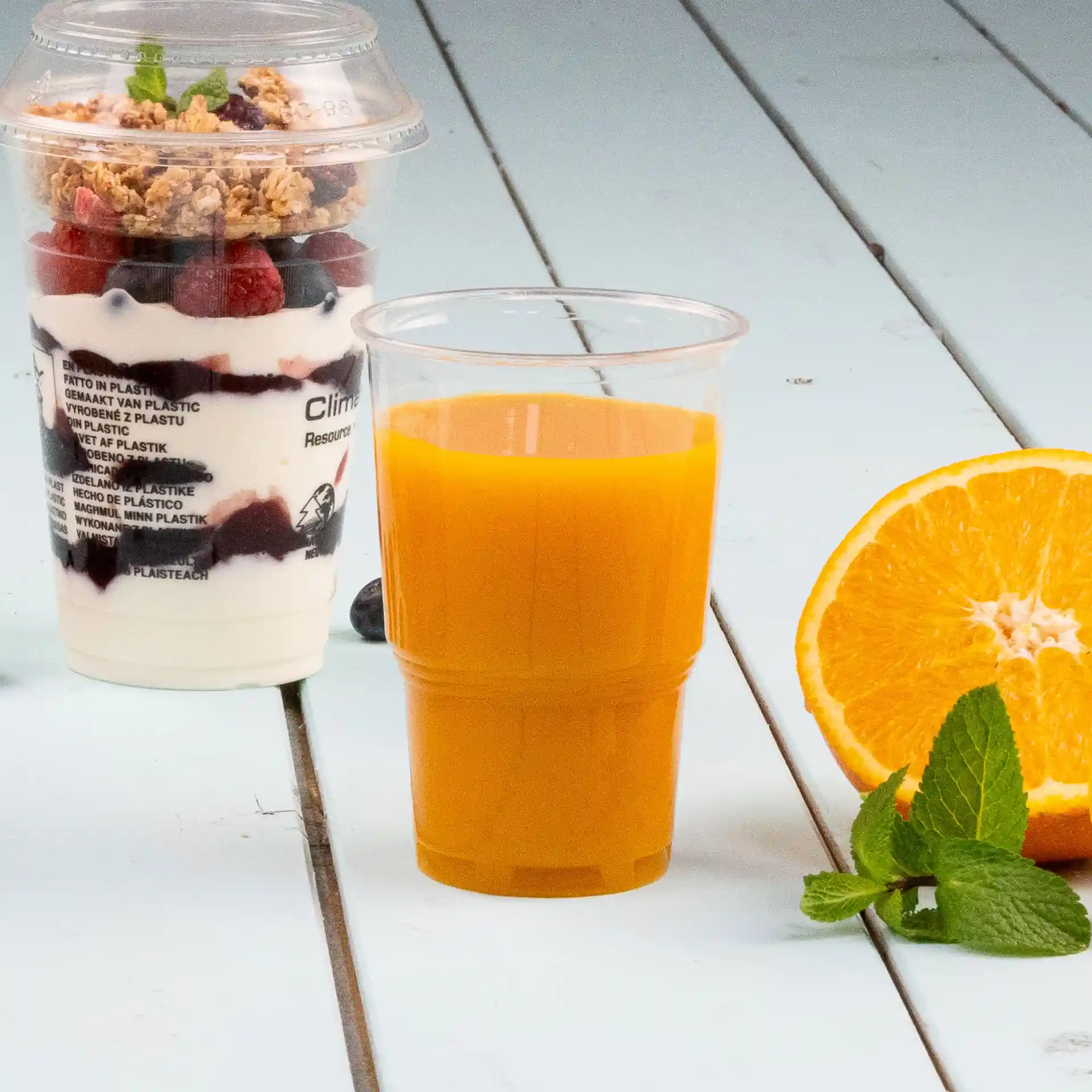 Immagine di un bicchiere di plastica trasparente riempito di succo d'arancia, accanto a un parfait di yogurt e una fetta d'arancia.