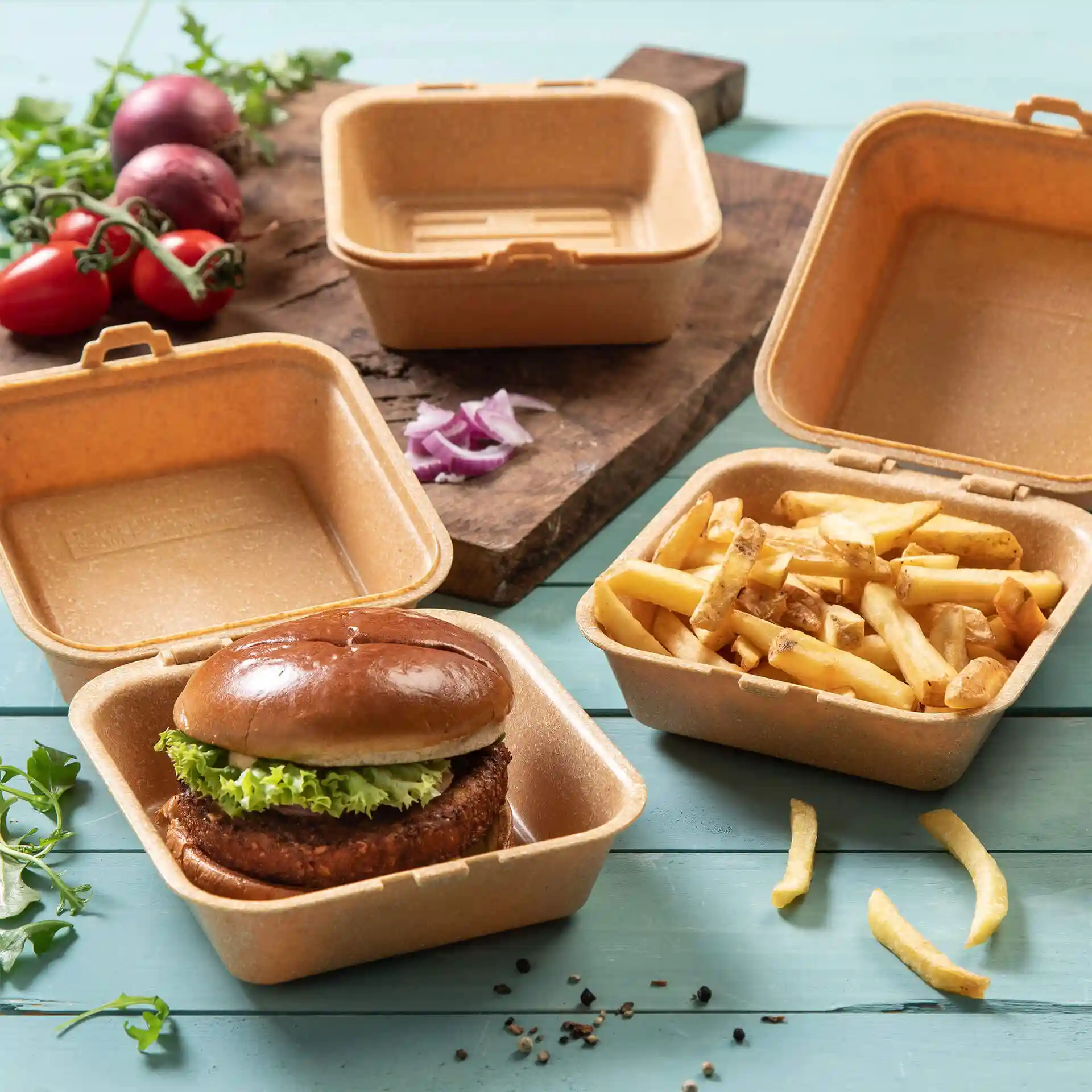 Scatola per hamburger riutilizzabile marrone con coperchio, contenente un hamburger e patatine, su una superficie di legno con verdure vicine.
