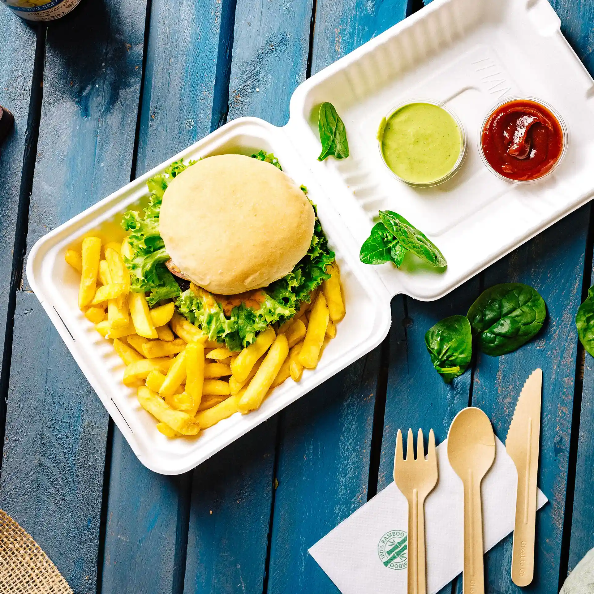 Contenitore rettangolare bianco con coperchio, con un hamburger, patatine, salsa verde, ketchup e posate di legno su una superficie blu.