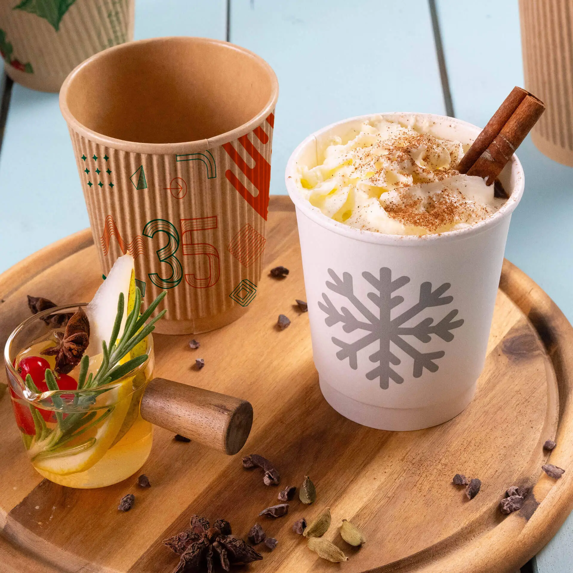 Un bicchierino di carta bianco con un design di fiocchi di neve, riempito di panna montata e un bastoncino di cannella; un bicchiere marrone a coste vicino.