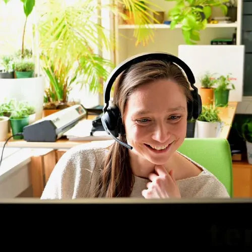 Donna con cuffie che sorride seduta a una scrivania con piante e un computer in una stanza luminosa.