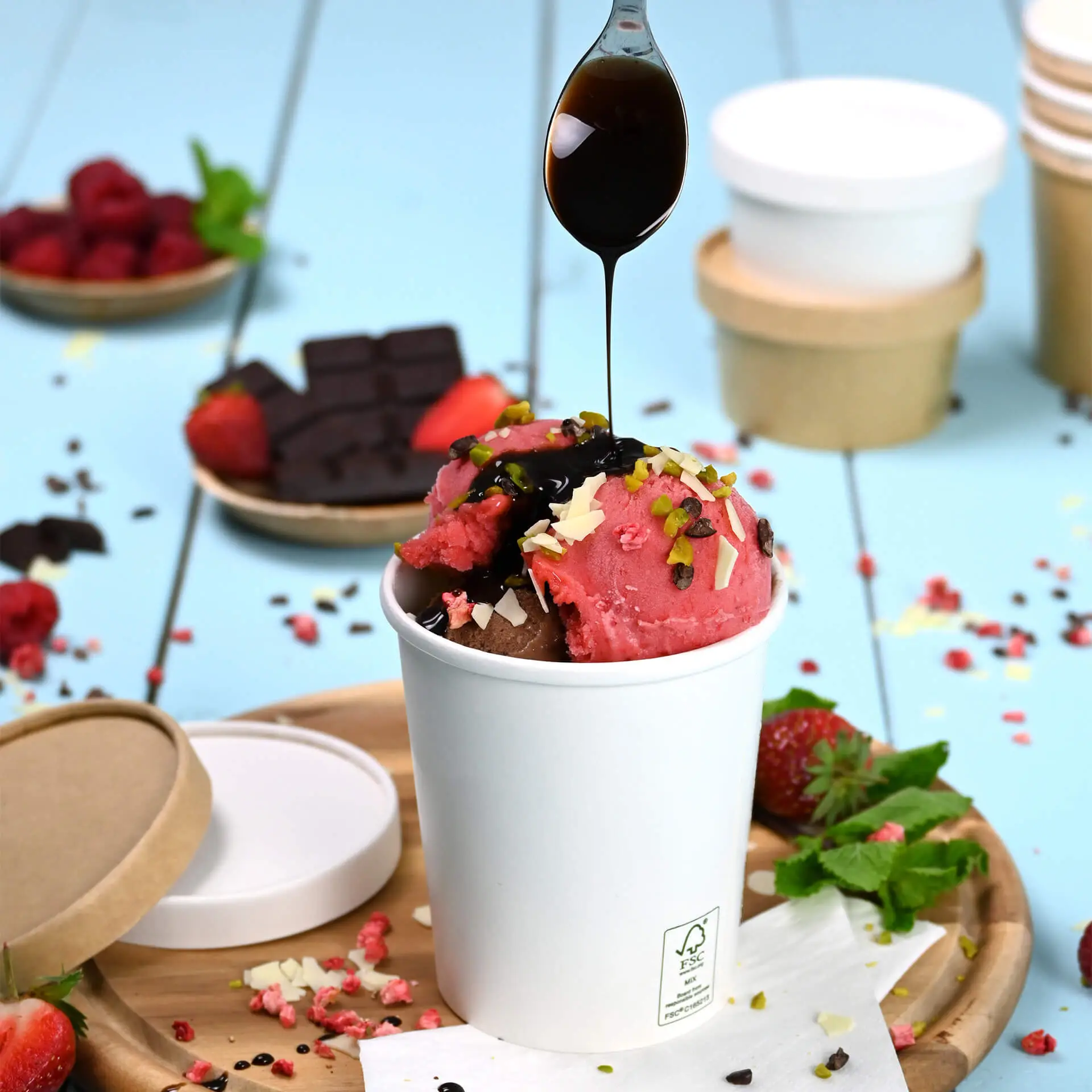 Coppetta di carta bianca riempita di gelato rosa e marrone, guarnita con salsa di cioccolato e noci, su un vassoio di legno.