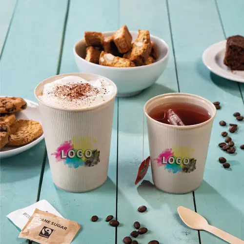 Due tazze con design del logo, una con panna montata e l'altra con tè freddo, circondate da biscotti, torta e pacchetti di zucchero.