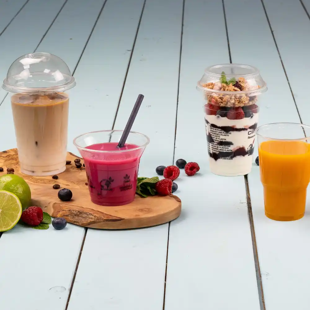 Quattro drink su un tagliere di legno: caffè freddo in un bicchiere con coperchio a cupola, bevanda rosa con cannuccia, bicchiere di dessert a strati e succo d'arancia.