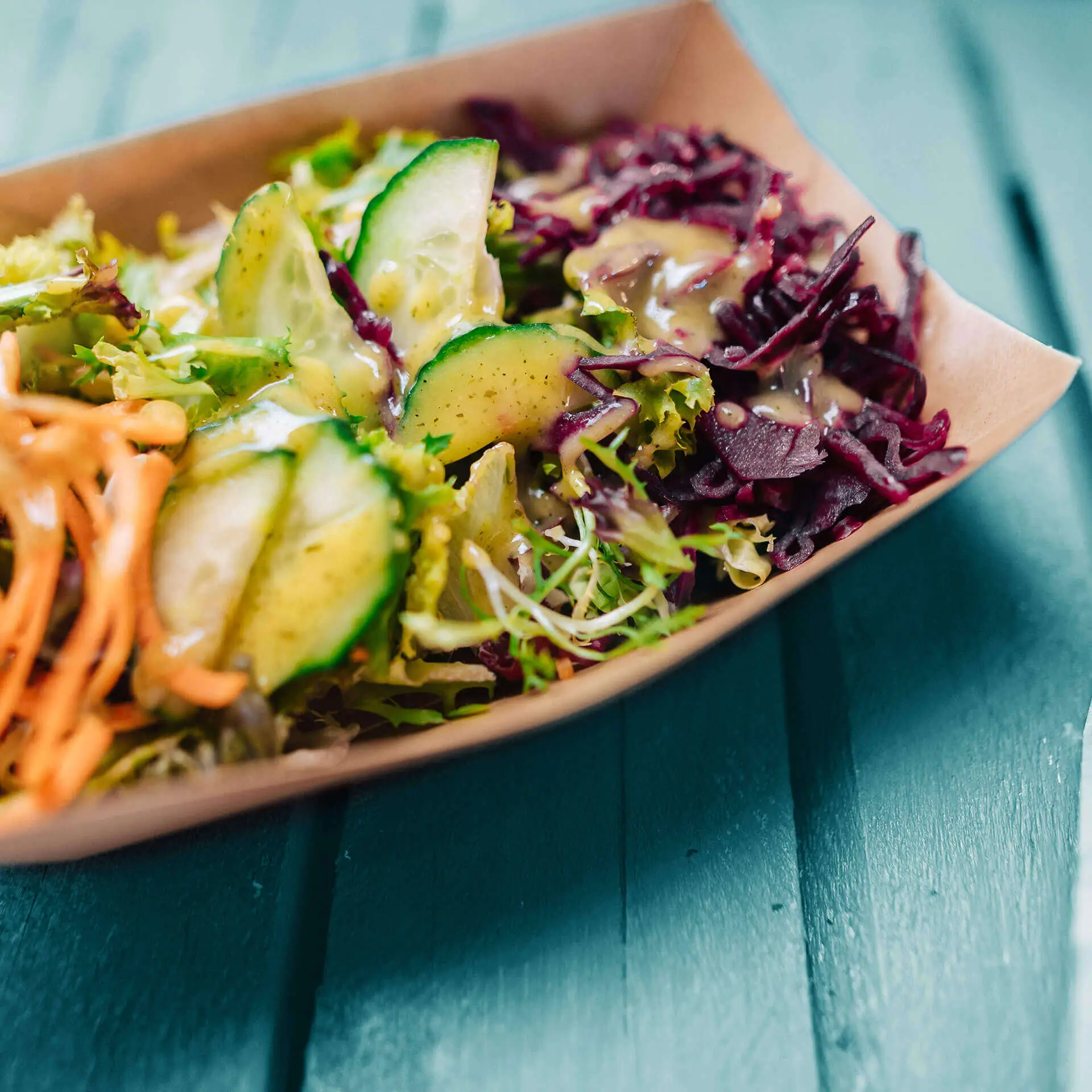 Una ciotola per snack piena di insalata mista, con fette di cetriolo e carote grattugiate su una superficie in legno verde.