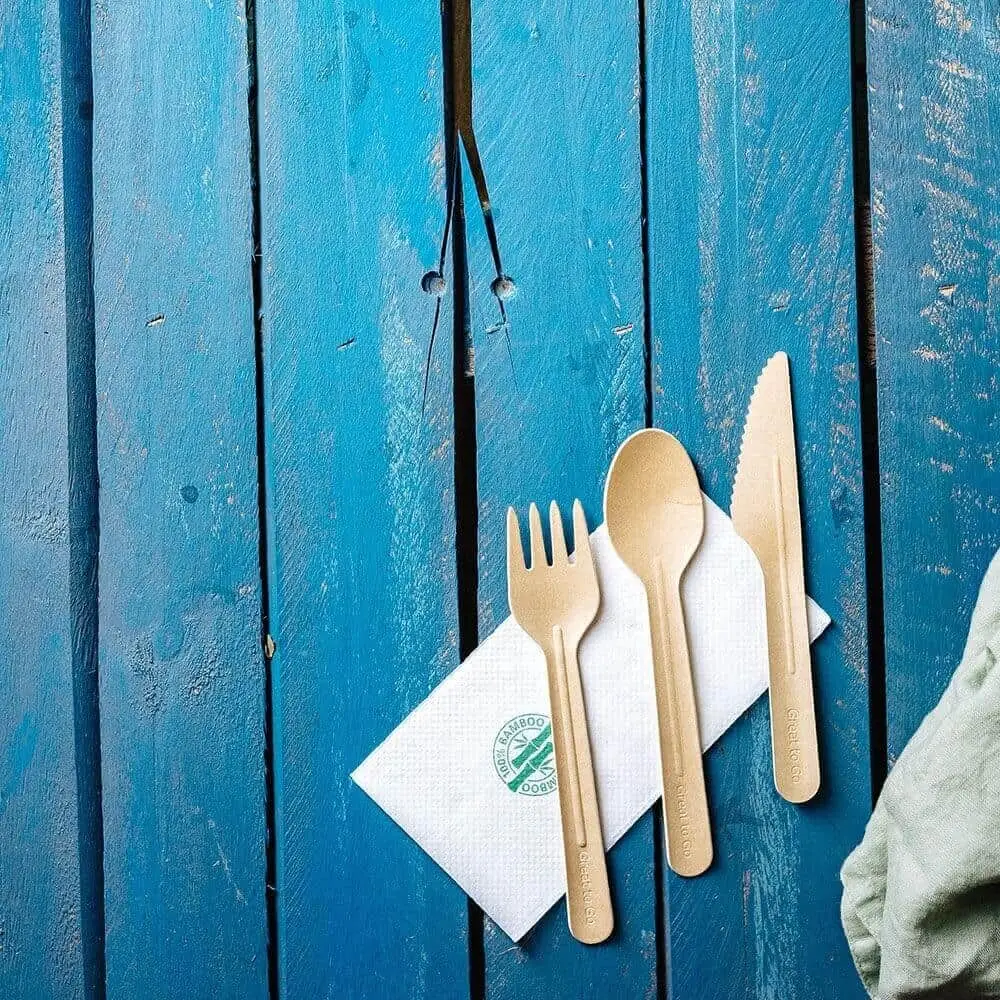 Un coltello, una forchetta e un cucchiaio di carta usa e getta su un tovagliolo bianco, posati su una superficie in legno blu.