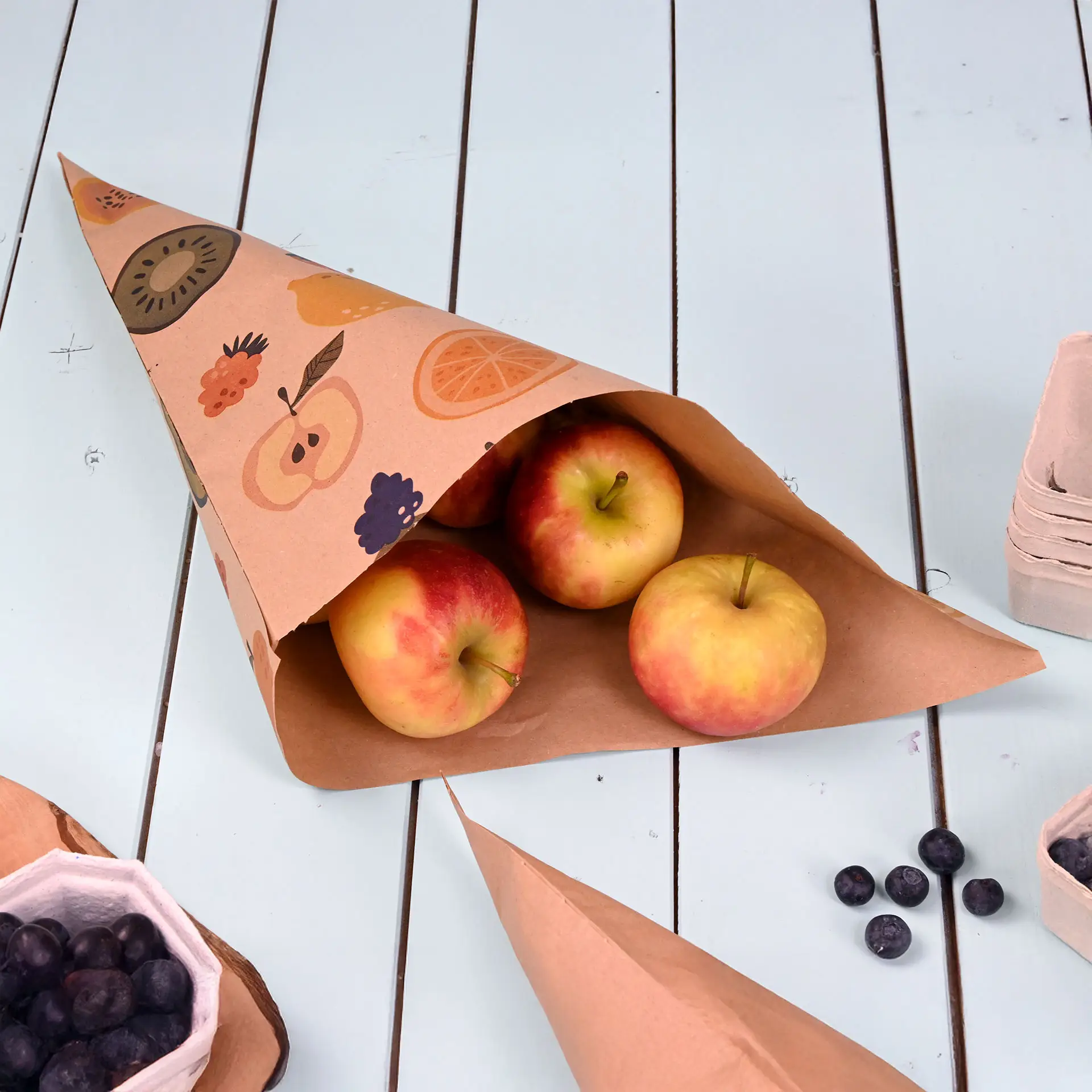 Cono di carta marrone con design di frutta, contenente tre mele rosse, su una superficie di tavolo azzurra.