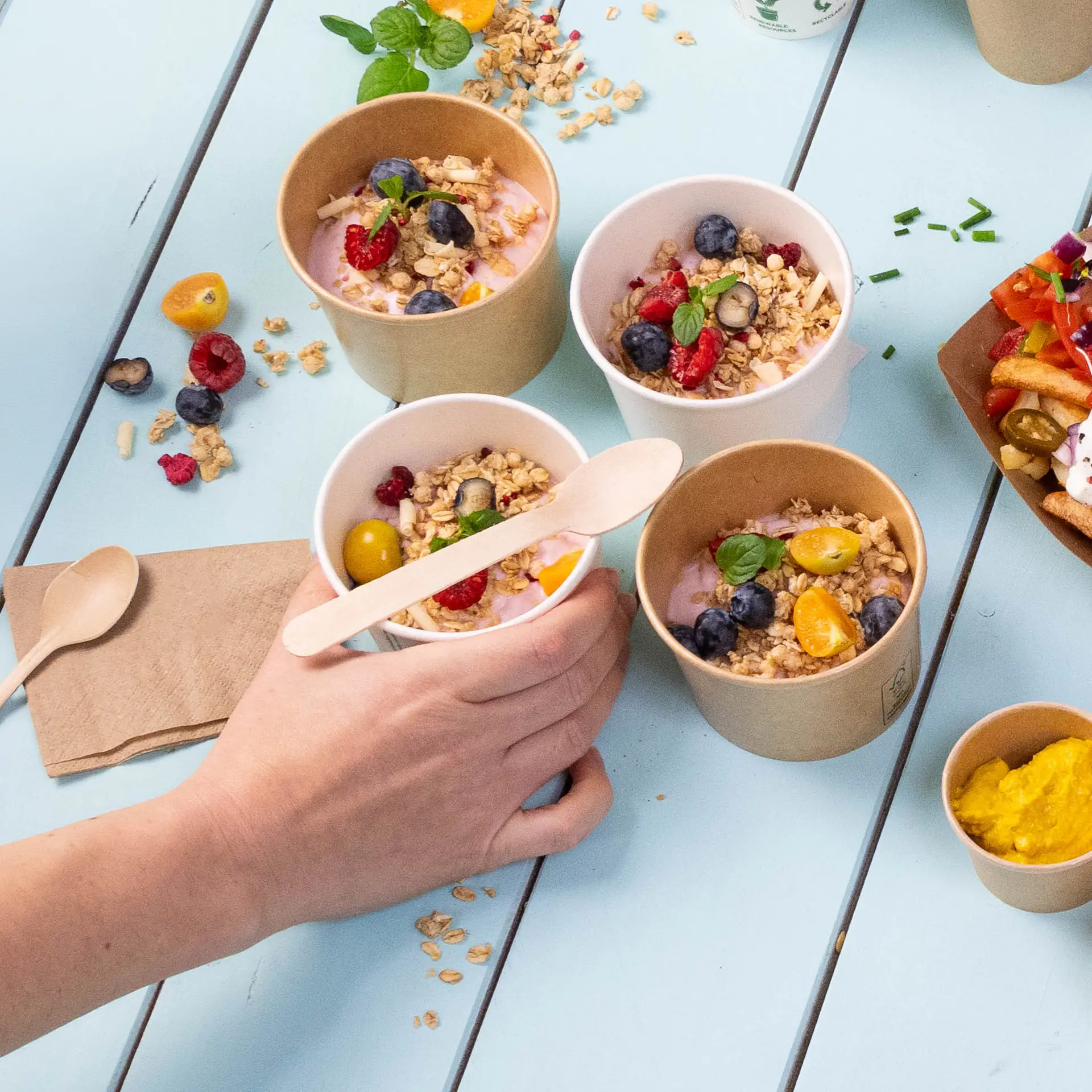 Immagine di diversi piccoli bicchieri con yogurt guarniti con granola e frutta, una mano che tiene un bicchiere e cucchiai di legno vicini.
