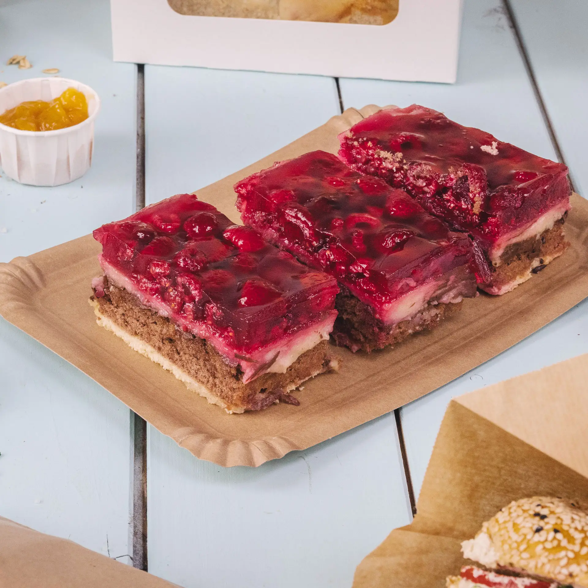 Quattro piatti rettangolari in cartone marrone con fette di dolce a strati guarnite con gelatina rossa e ciliegie.