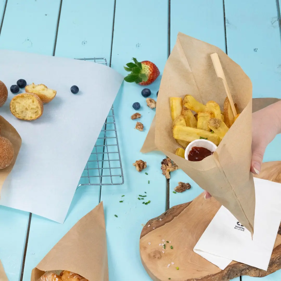 Cono di carta marrone riempito di patatine e un piccolo bicchiere di ketchup, circondato da dolci, frutti di bosco e un tagliere di legno.