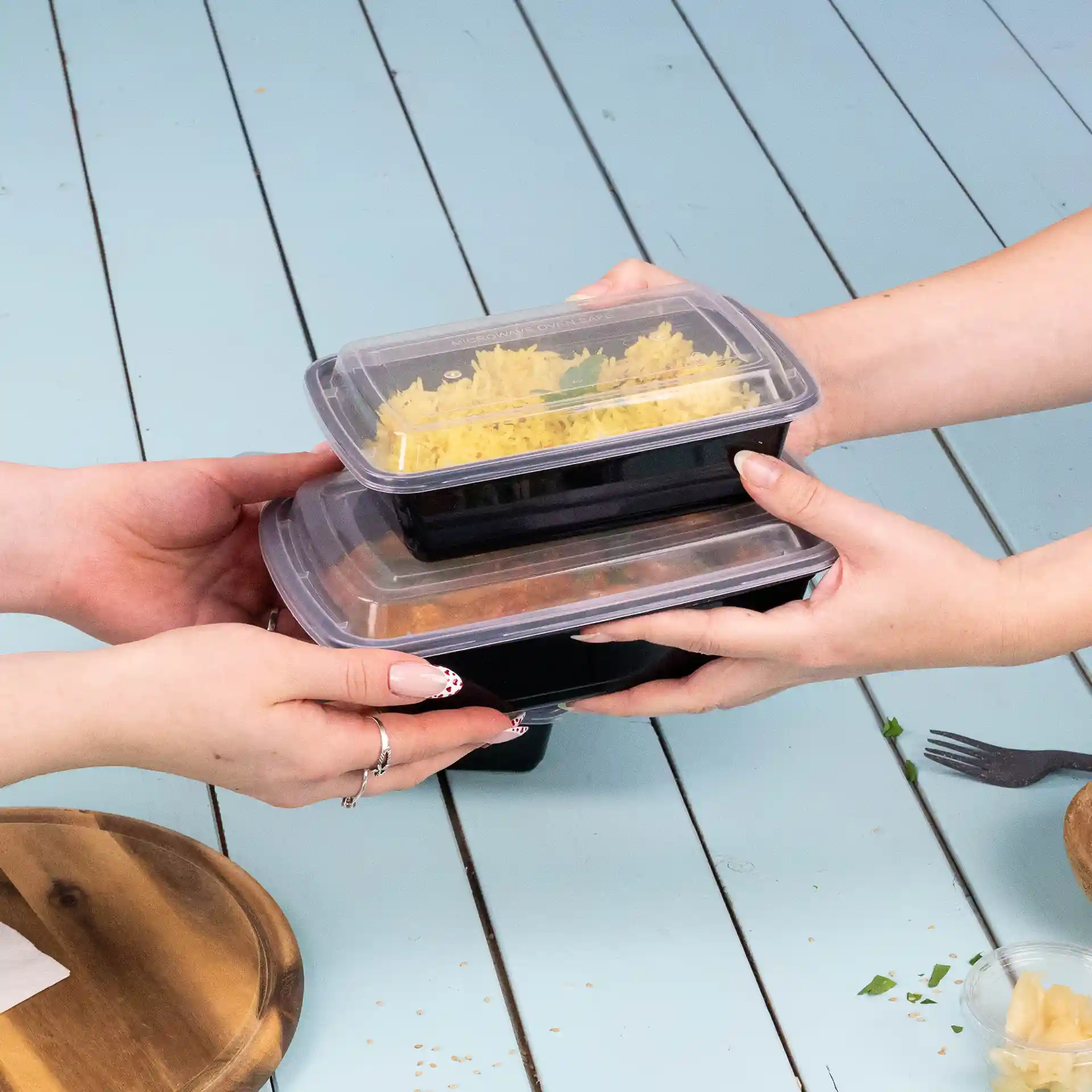 Due mani tengono contenitori alimentari rettangolari impilati con coperchi trasparenti, su una superficie azzurra.