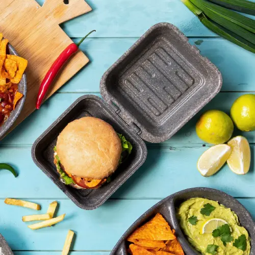 Burger in una scatola di kraft riutilizzabile con lattuga e salse, circondato da patatine, guacamole e spicchi di lime.