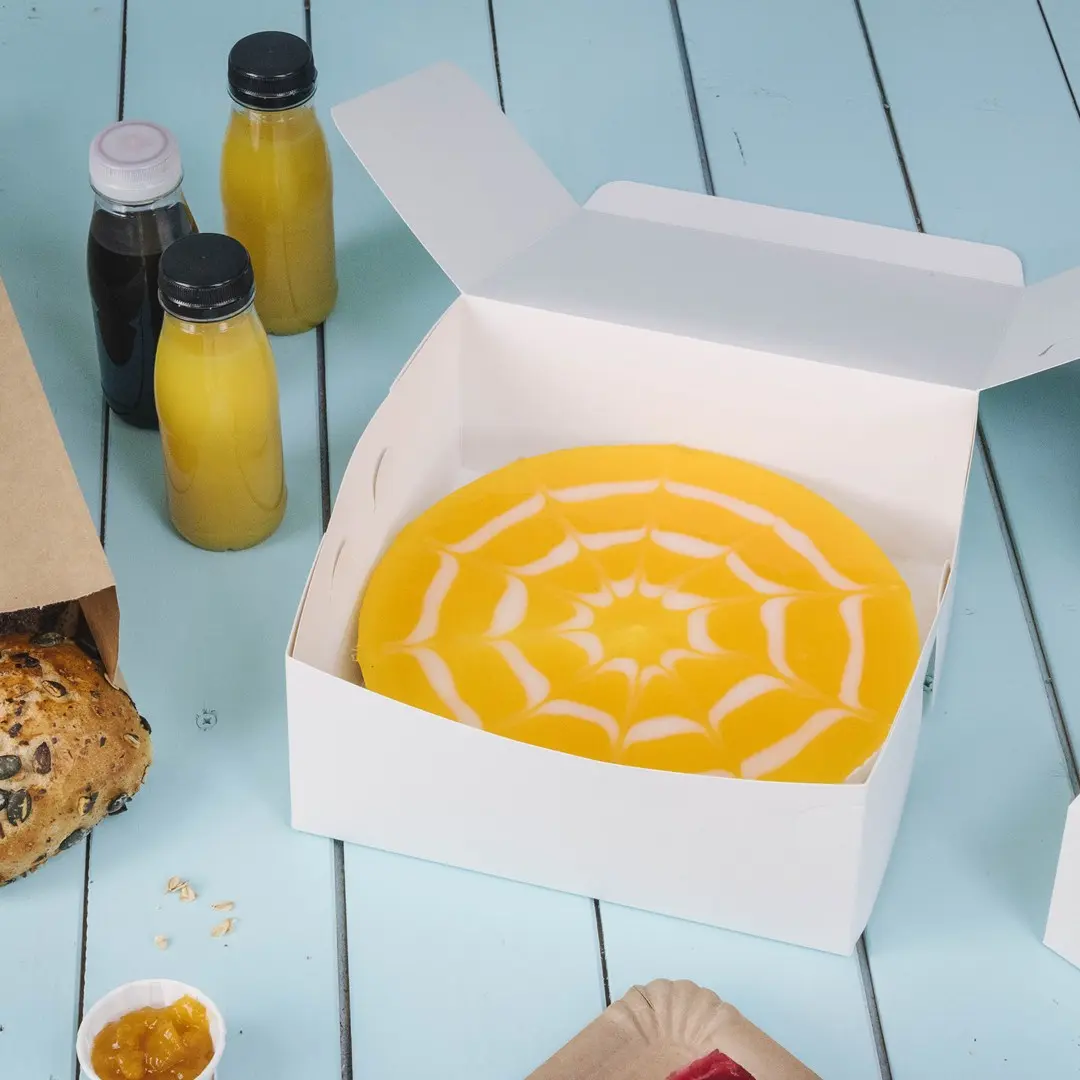Scatola bianca per dolci contenente una torta gialla con un design di ragnatela bianca, circondata da bevande e prodotti da forno.