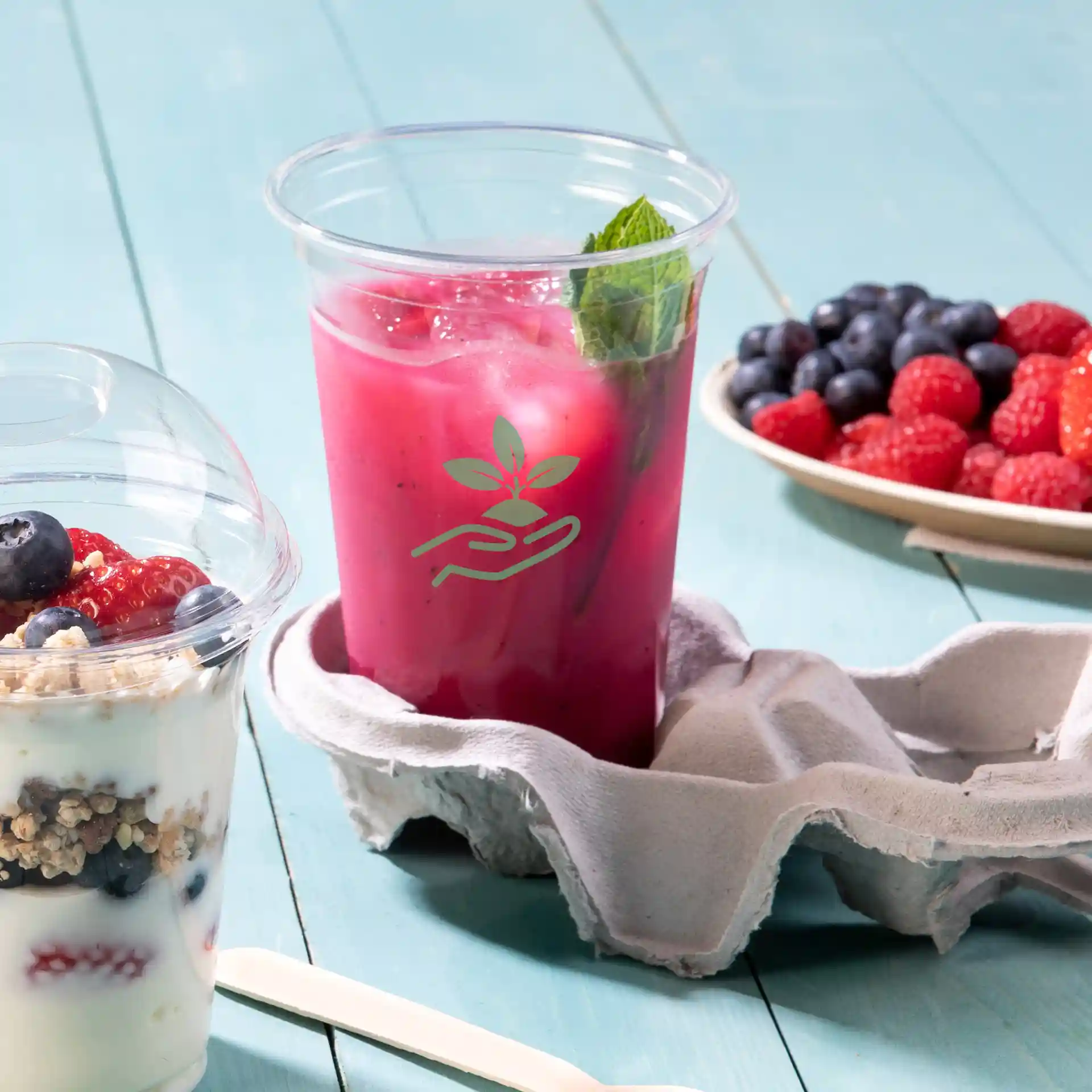 Bicchiere rPET trasparente da 16 oz riempito con bevanda rosa e menta, posizionato in un portauova con un parfait di yogurt vicino.