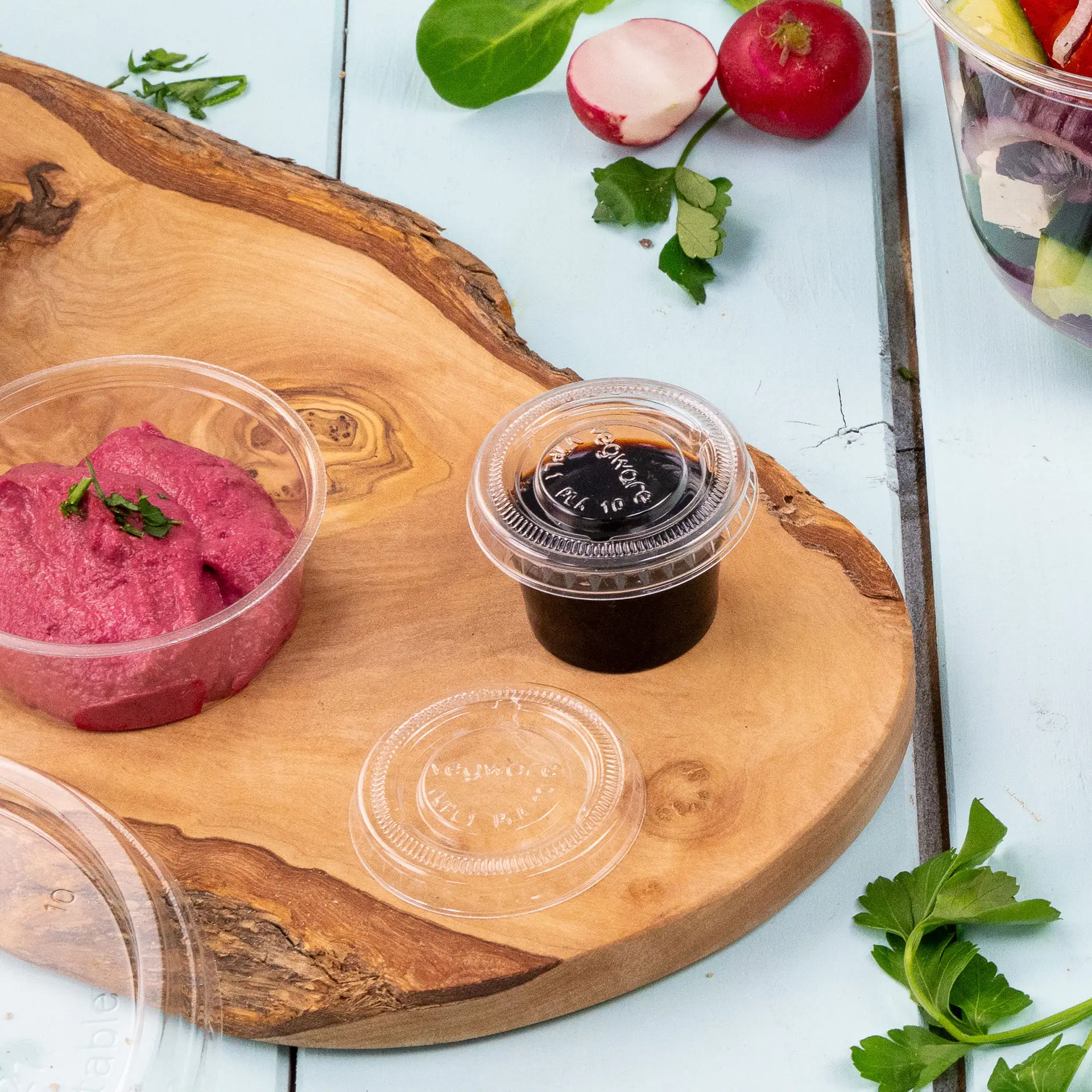 Un coperchio trasparente e due piccoli contenitori: uno con salsa rosa e uno con salsa scura, su una superficie di legno.