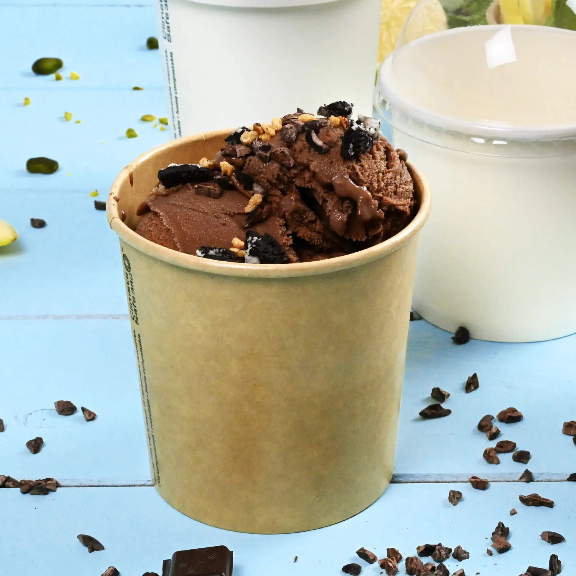 Una coppa di carta marrone riempita di gelato al cioccolato, guarnita con biscotti sbriciolati e noci, circondata da piccoli pezzi di cioccolato.