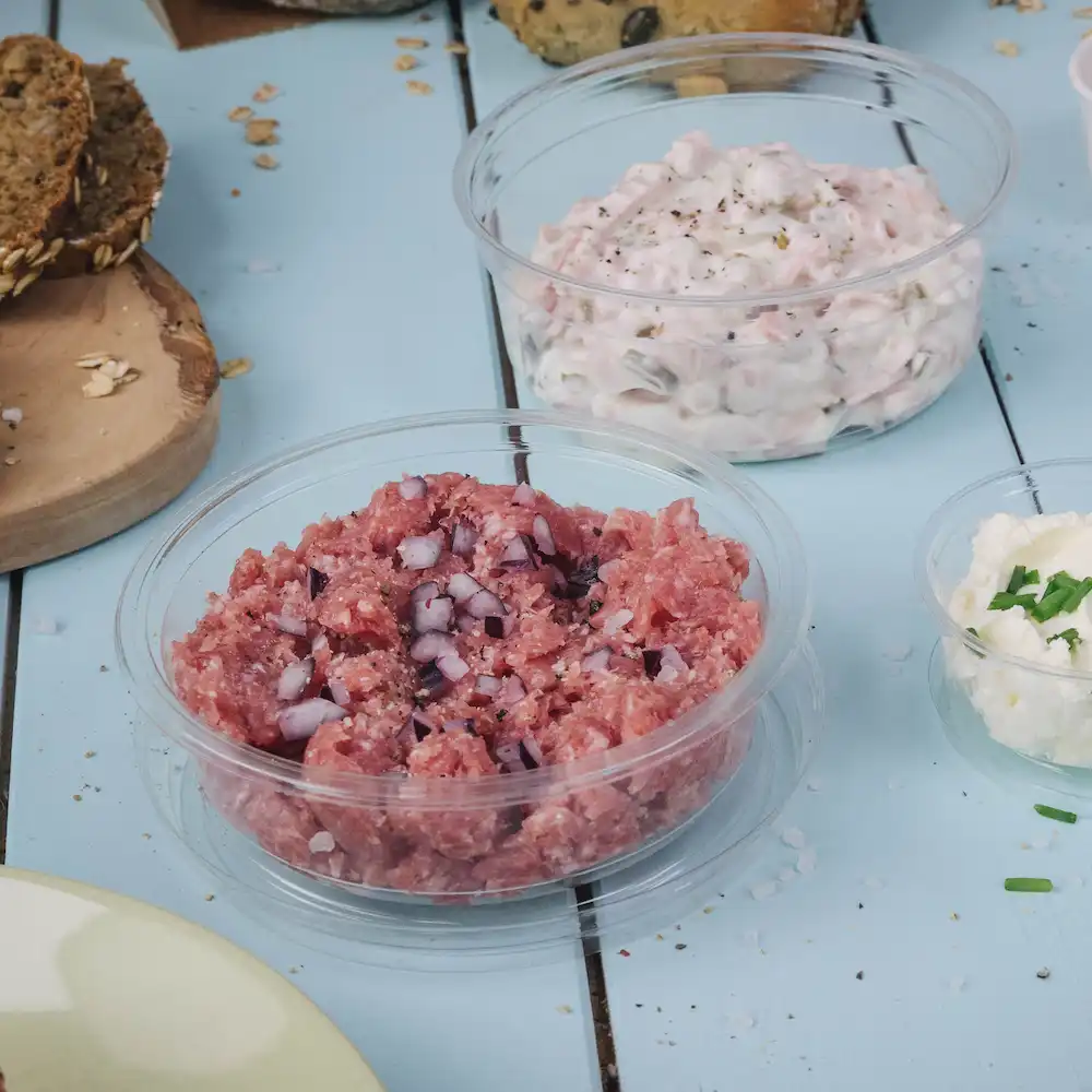 Carne cruda tritata con cipolle a dadini in un contenitore trasparente; accanto, salsa cremosa in un altro contenitore e una ciotola di formaggio.