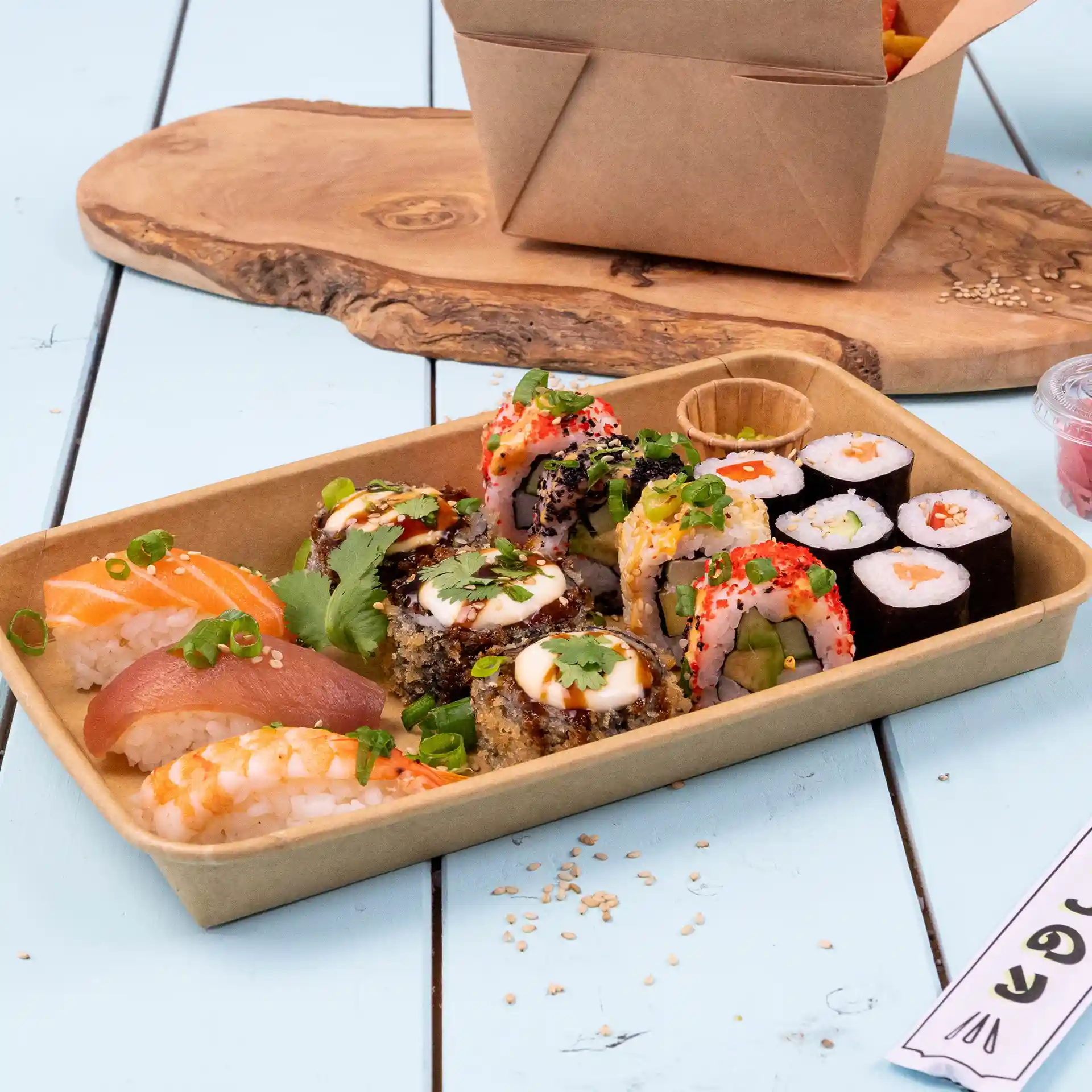 Assortimento di sushi in una scatola marrone rivestita in PE, con vari rotoli e sashimi, su una superficie di legno.
