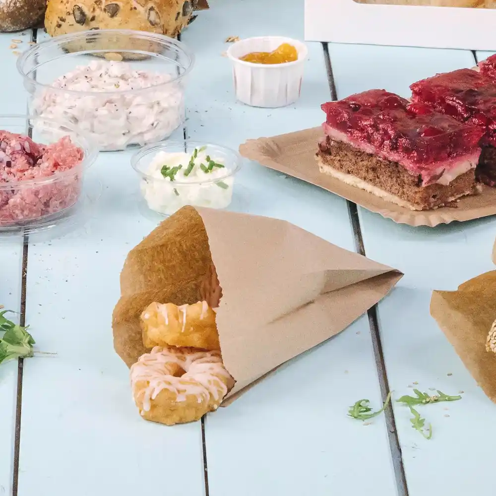 Sacchetto di carta marrone contenente due donut ghiacciati, circondato da vari alimenti e guarnizioni su un tavolo blu.