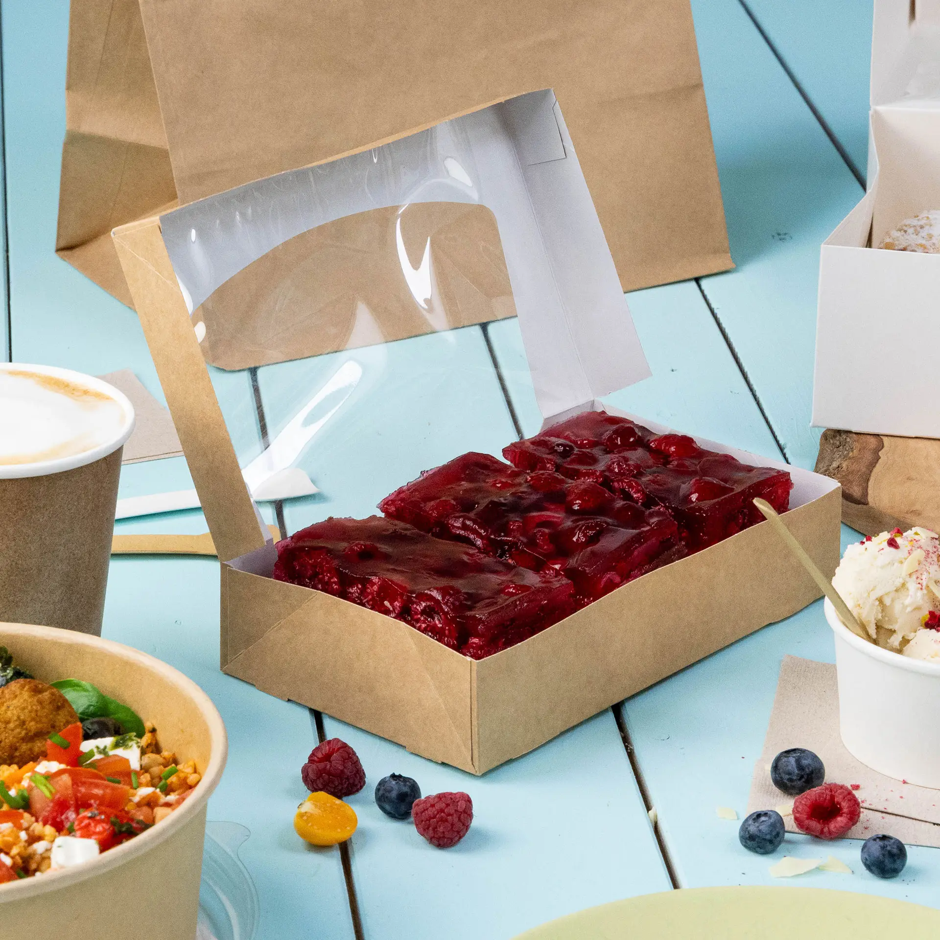 Una scatola pieghevole in cartone con un coperchio in cellulosa trasparente, contenente dolci rossi, su uno sfondo blu con altri alimenti.