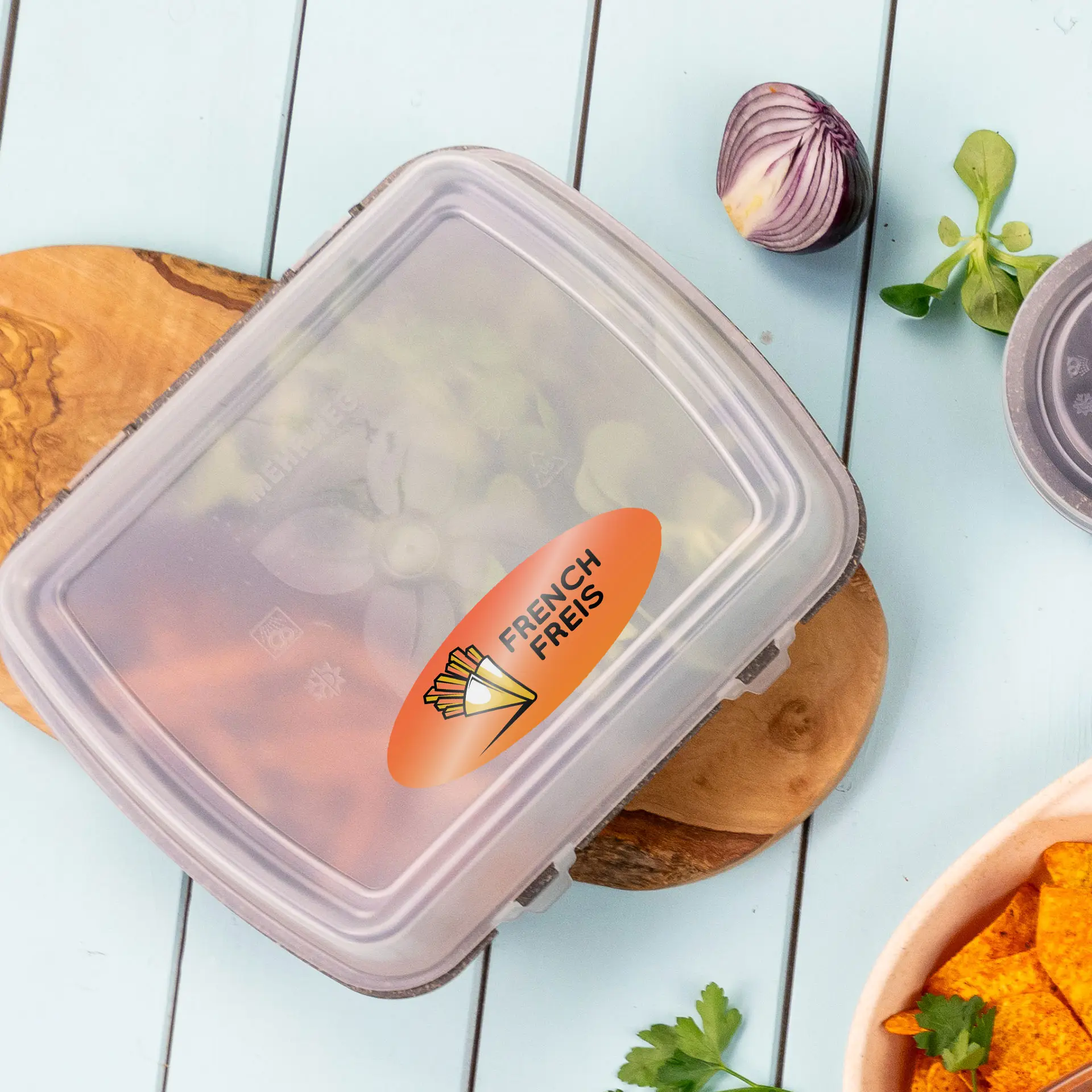 Etichetta ovale trasparente con il testo "FRENCH FRIES" su un contenitore, circondata da ingredienti freschi su un tagliere di legno.