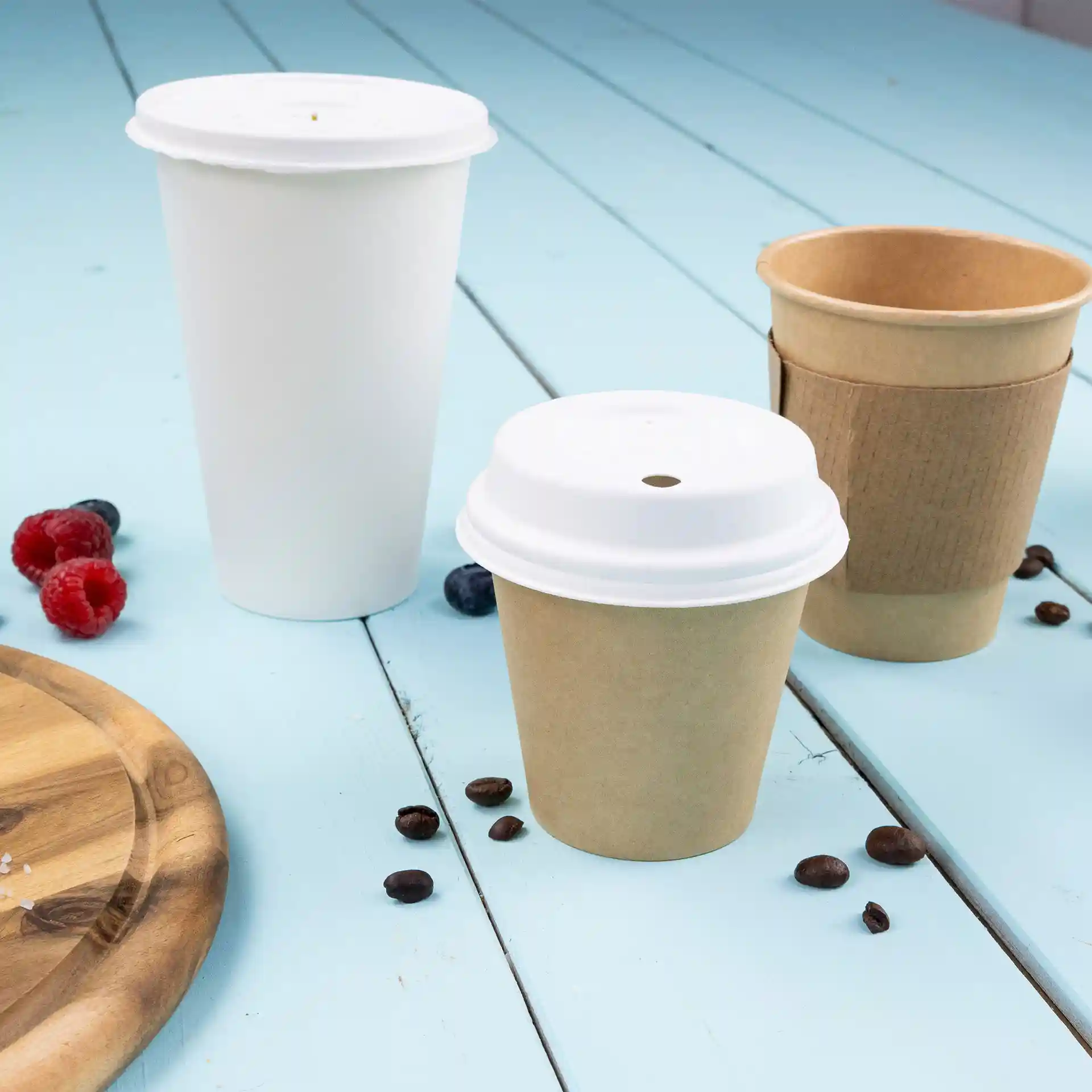 Immagine di tre bicchieri di carta: uno bianco, uno marrone con un coperchio bianco e uno marrone senza coperchio, su una superficie blu con chicchi di caffè e bacche.