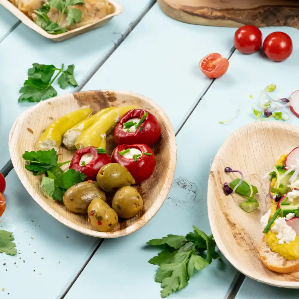 Un piatto di legno con peperoni rossi ripieni, peperoni gialli sott'aceto e olive verdi; erbe fresche sparse su una superficie blu.