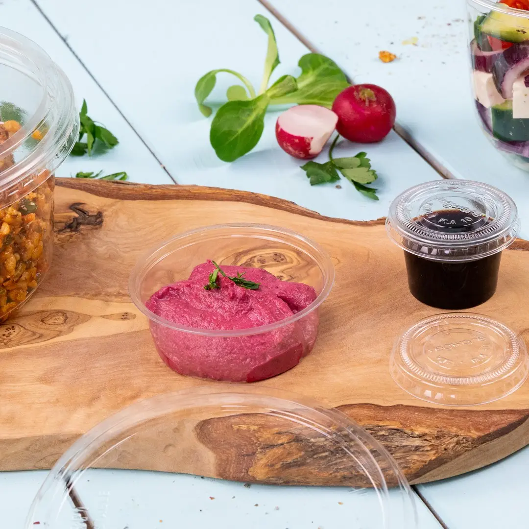 Bicchiere di plastica trasparente con salsa rosa, circondato da un tagliere di legno, ingredienti per insalata e un altro piccolo bicchiere.