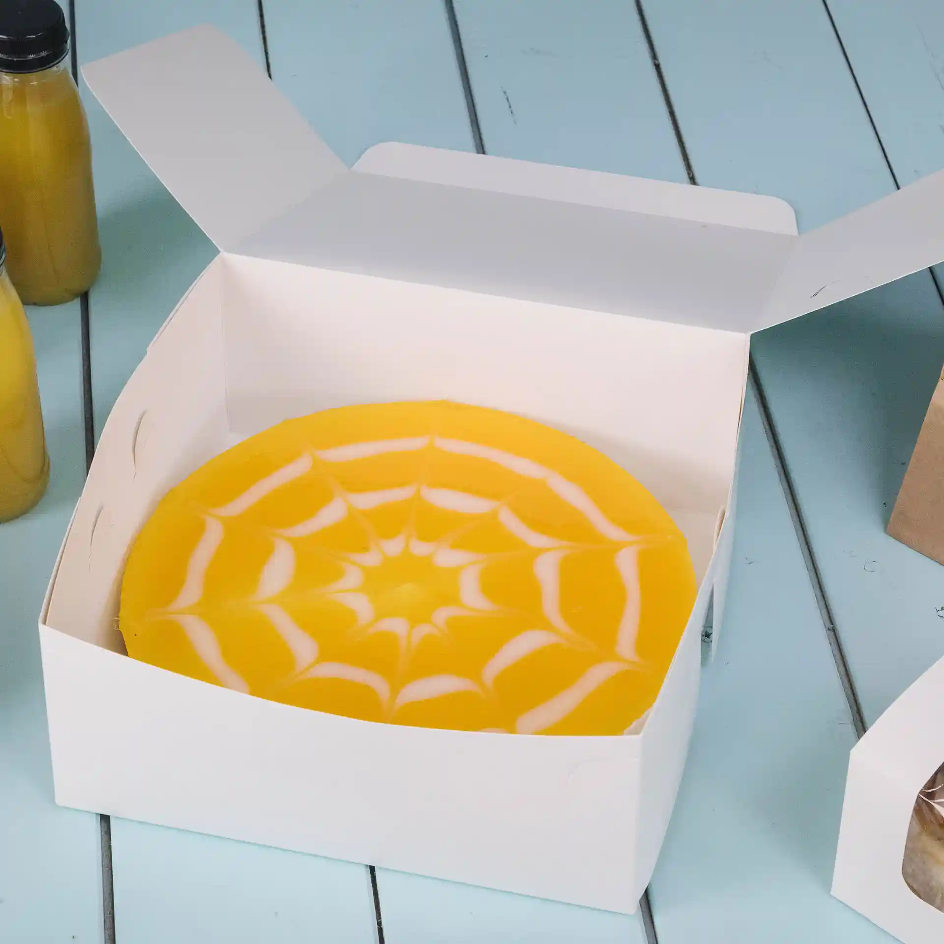 Scatola per dolci bianca con una torta gialla con design di ragnatela all'interno, posizionata su una superficie blu.