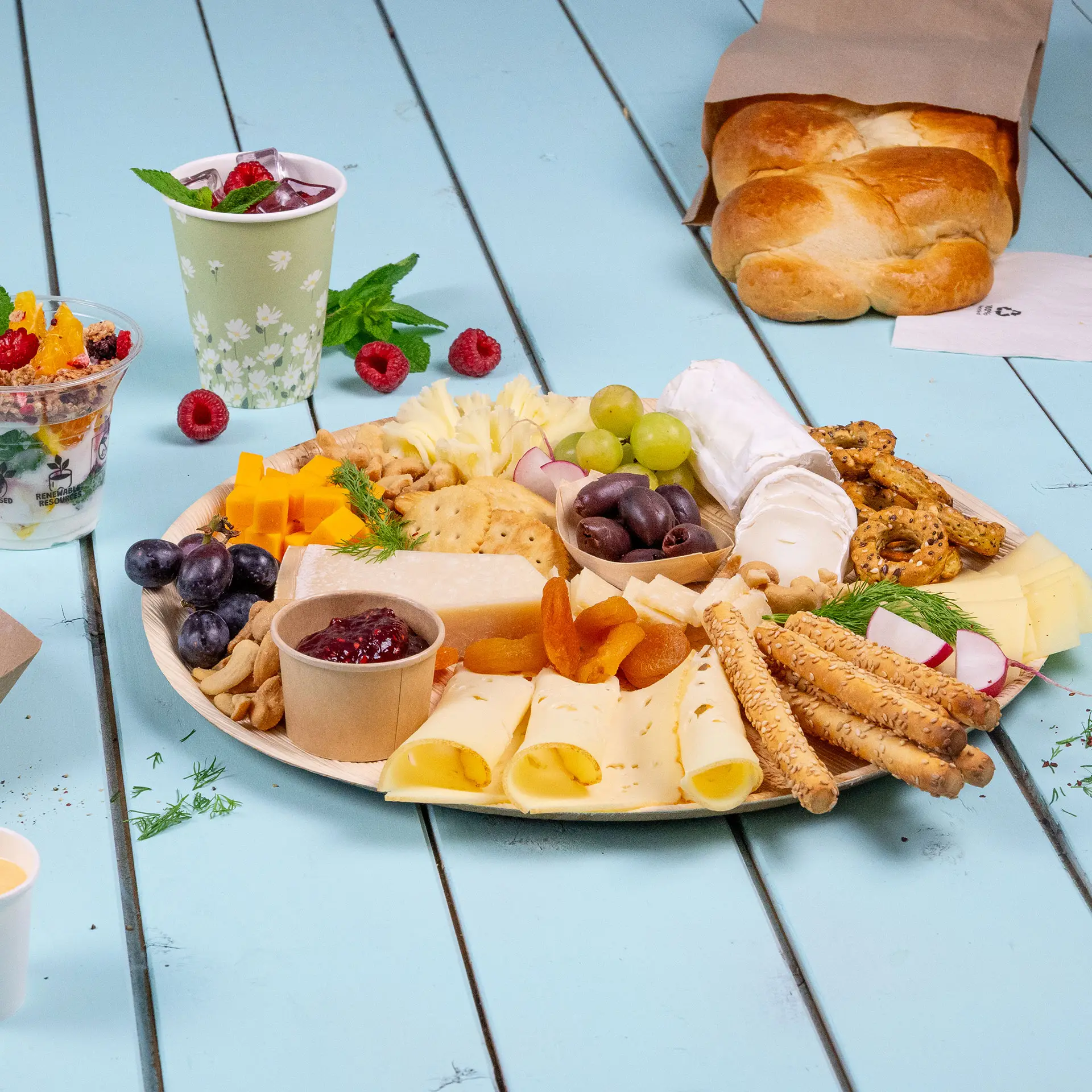 Un piatto rotondo in foglia di palma con una selezione di formaggi, frutta, noci e cracker, insieme a un pane e bevande.