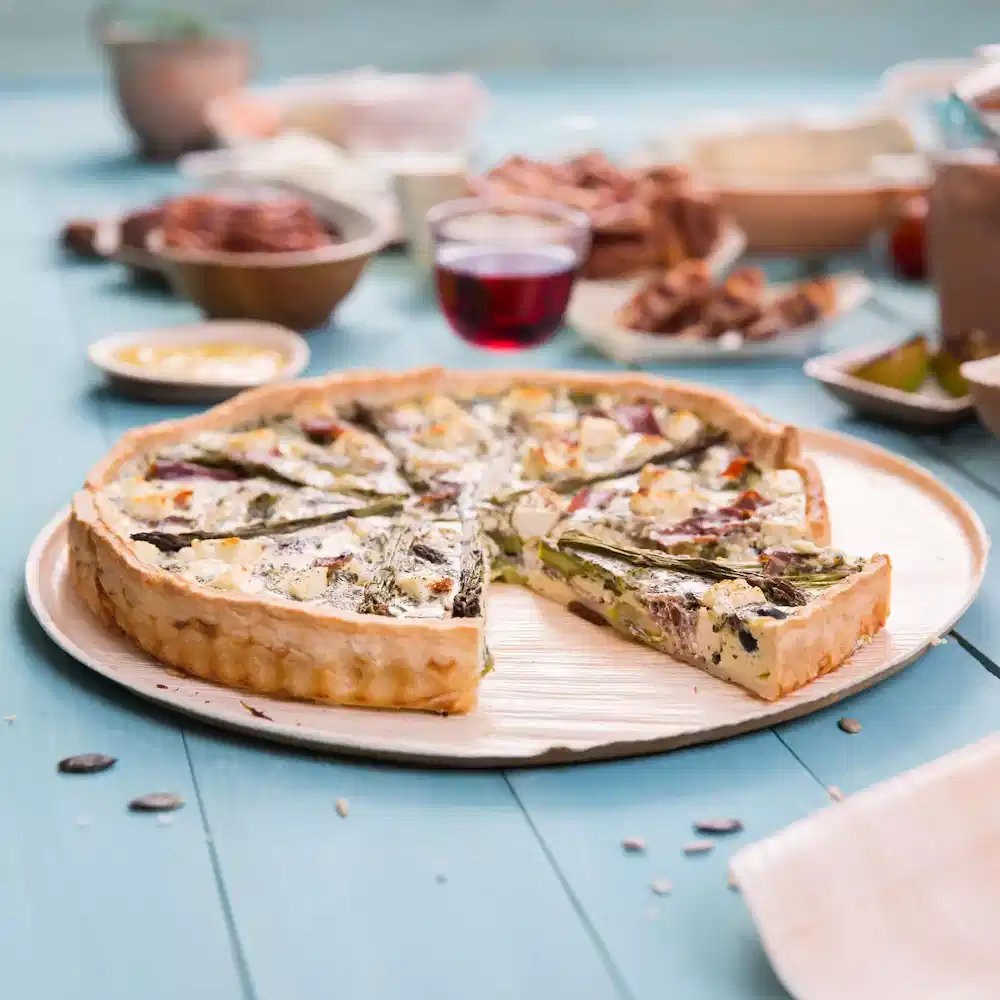 Quiche salata su un piatto di legno, con una fetta rimossa, circondata da vari piatti e un bicchiere di bevanda rossa.