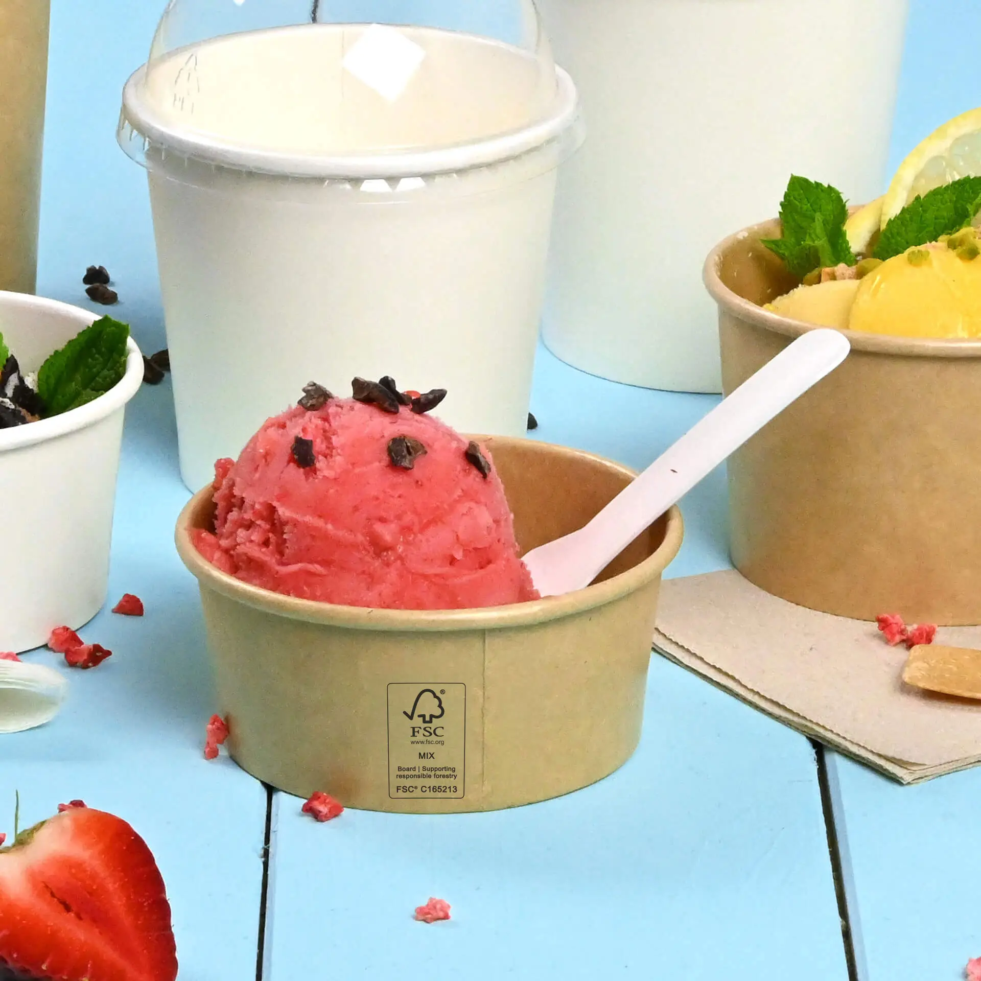 Bicchiere di carta marrone con gelato rosa e gocce di cioccolato, cucchiaio bianco e altri bicchieri sullo sfondo.