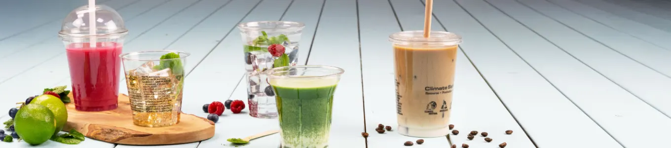 Bevande assortite esposte su una superficie di legno, tra cui caffè freddo, smoothie, parfait di yogurt e succo, con una fetta d'arancia accanto.