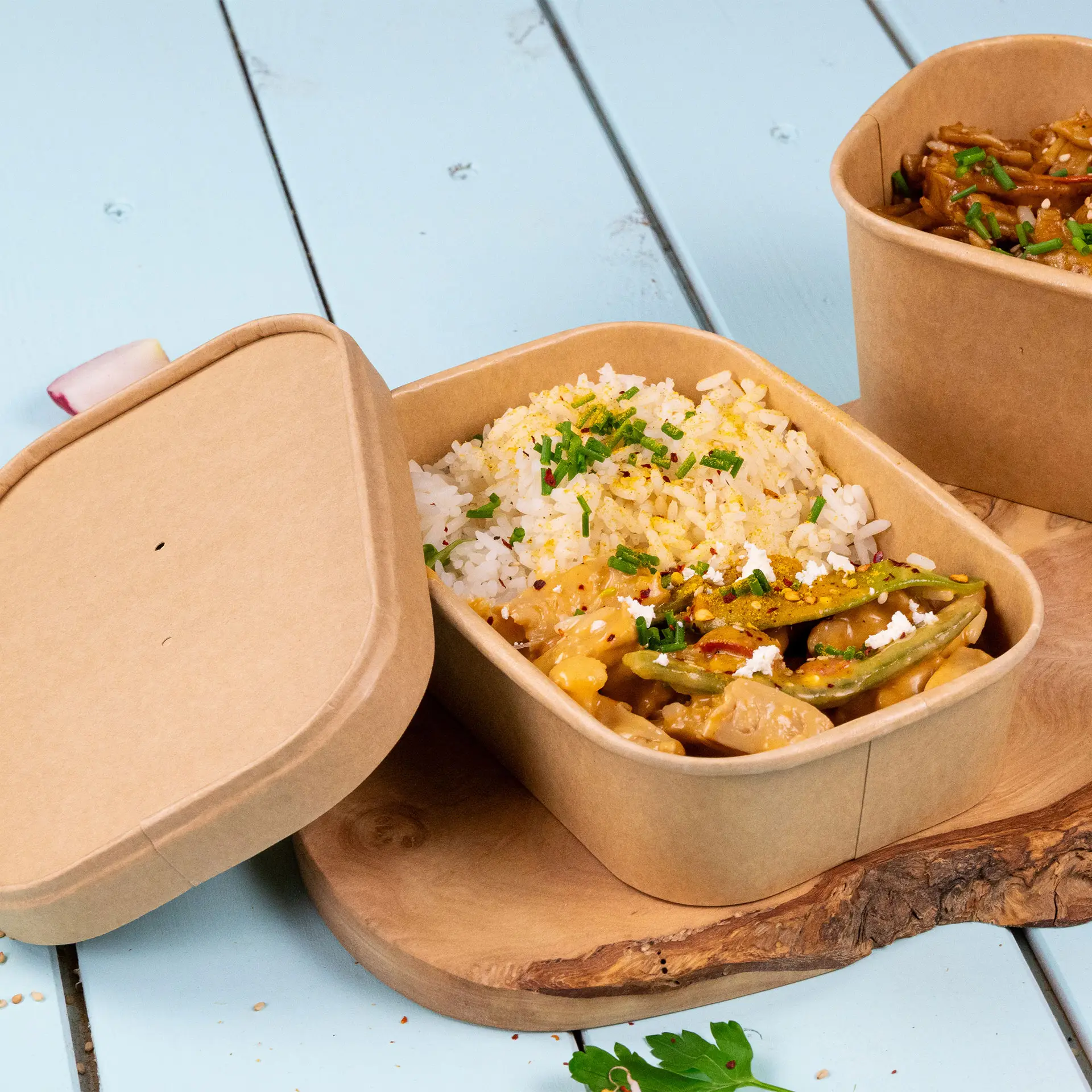 Contenitore alimentare rettangolare in kraft con riso e verdure, leggermente aperto; mostrato su un tagliere di legno con un contenitore simile accanto.