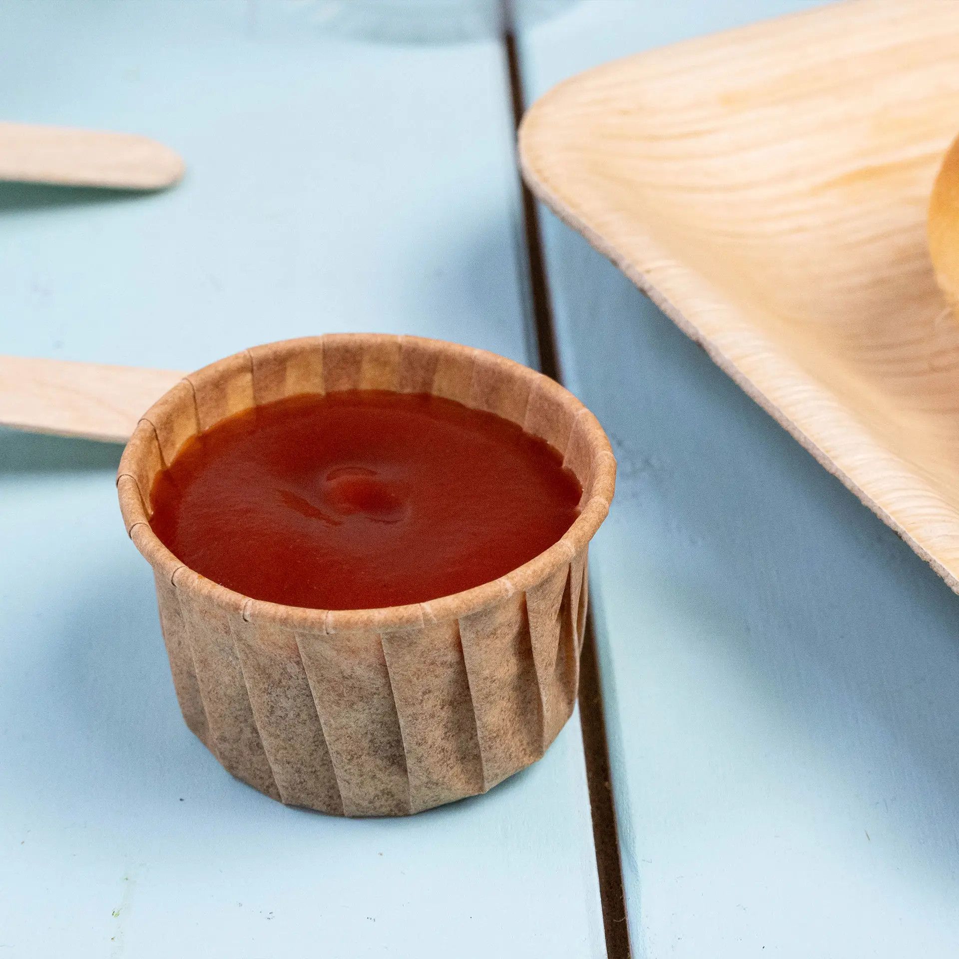 Coppetta di carta marrone riempita di ketchup su una superficie blu, progettata per salse e piccole porzioni.