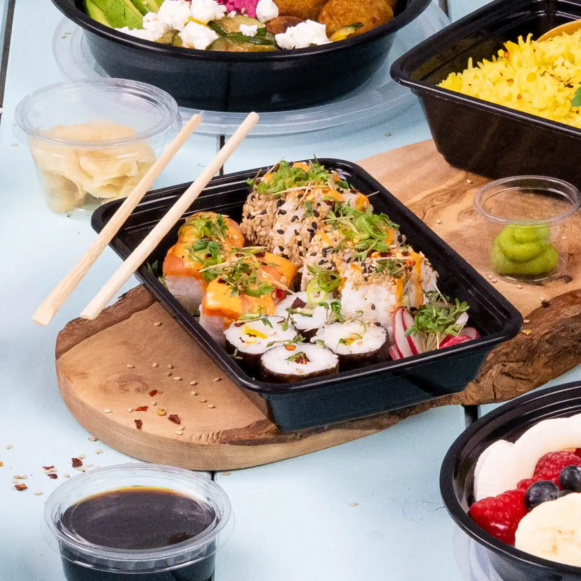 Rolls di sushi con condimenti in un contenitore rettangolare nero riutilizzabile, bacchette accanto e condimenti tra cui wasabi e salsa di soia.
