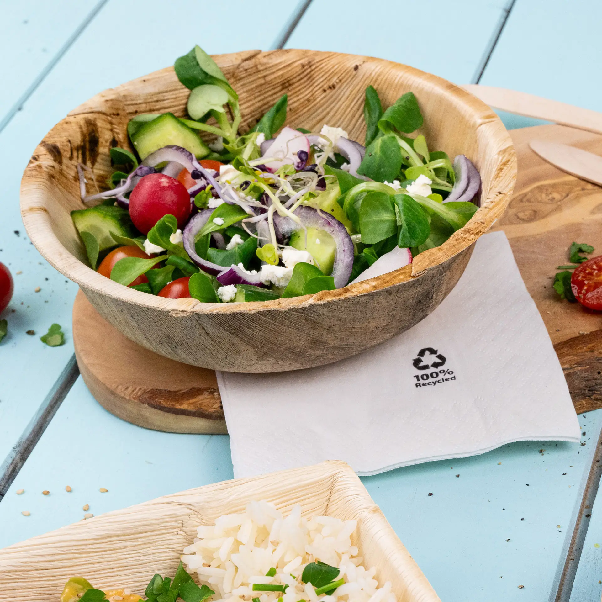 Una ciotola rotonda di foglie di palma riempita di ingredienti per insalata mista, posata su un tagliere di legno accanto a un tovagliolo contrassegnato con simboli di riciclo.