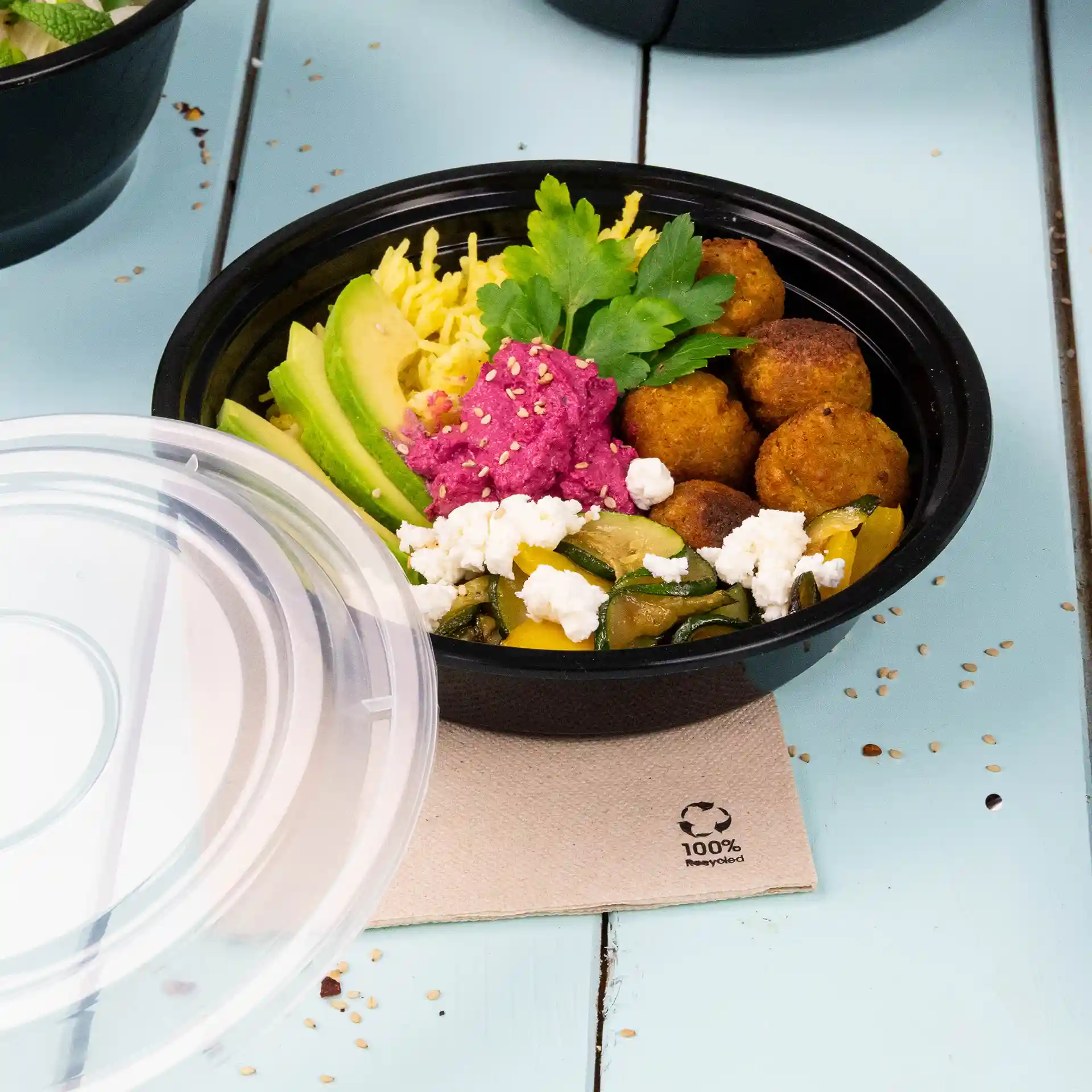 Piatto rotondo riutilizzabile nero con vari alimenti, tra cui avocado, falafel e una crema rosa, su una superficie azzurro chiaro.
