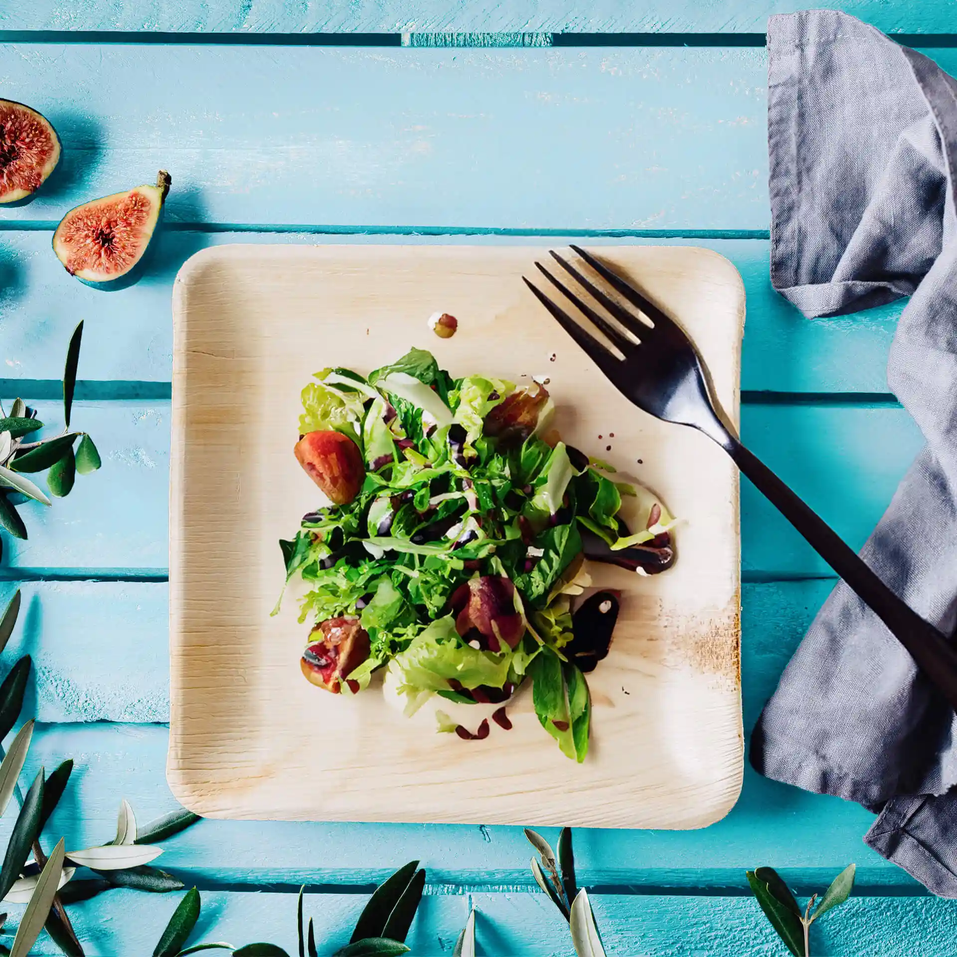 Piatto quadrato in foglia di palma con insalata guarnita con verdure e fichi, accompagnato da una forchetta nera su una superficie in legno blu.